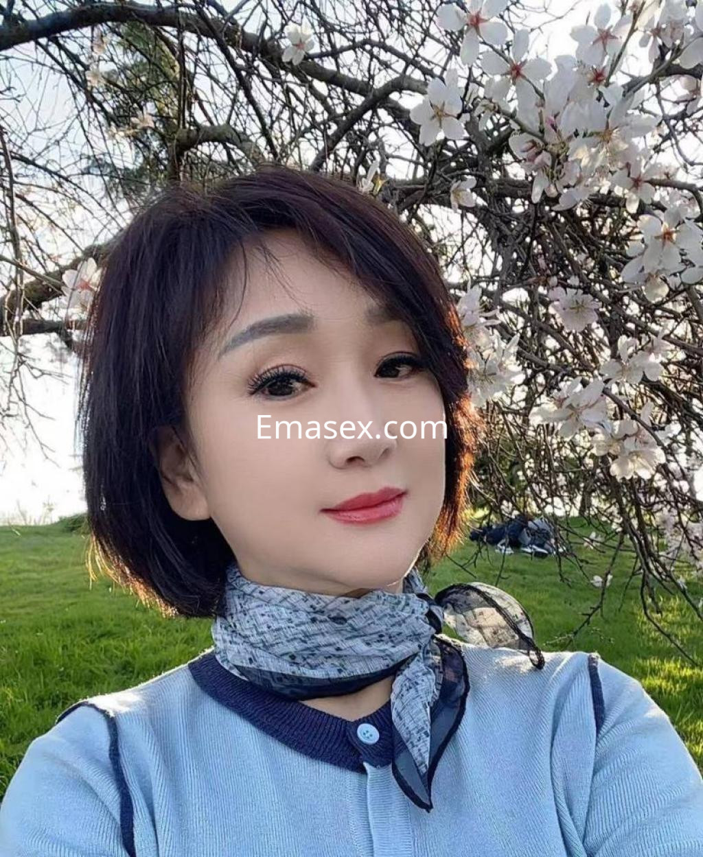 Escort en Barcelona con cabello corto y lacio, luciendo un suéter azul y una bufanda, posando entre flores.