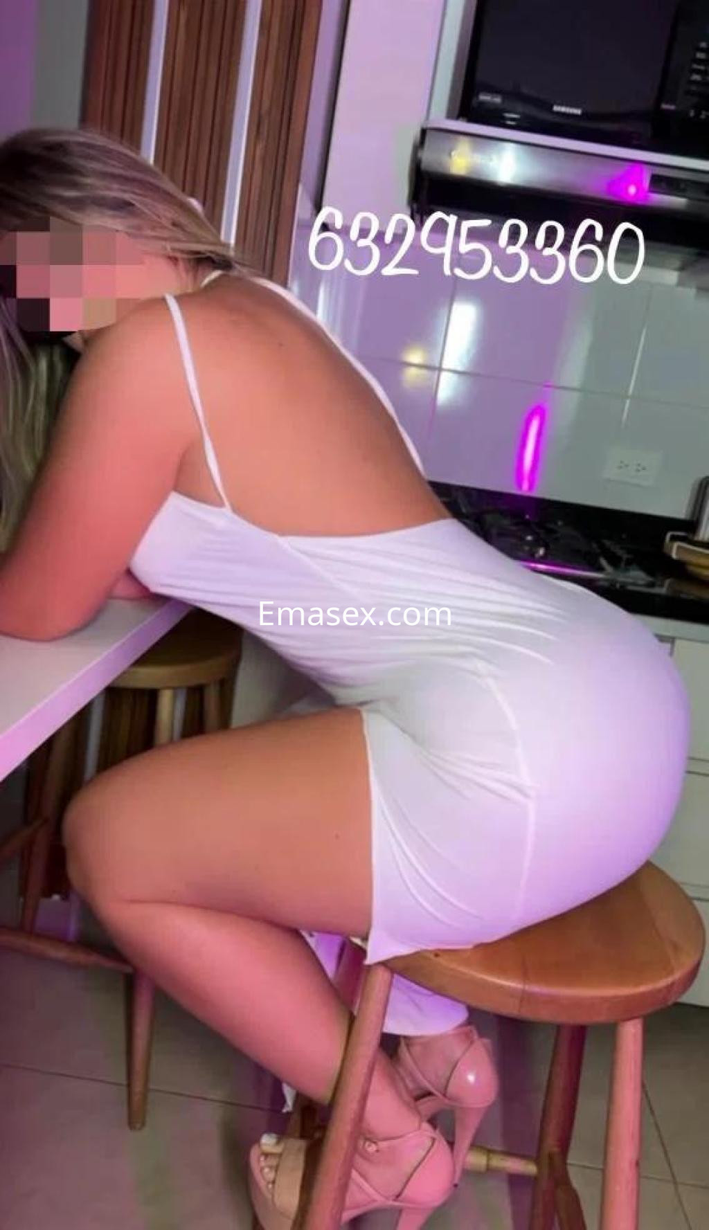 Escort en Coruña, con vestido blanco de tirantes, posando de lado sobre una silla en una cocina moderna.