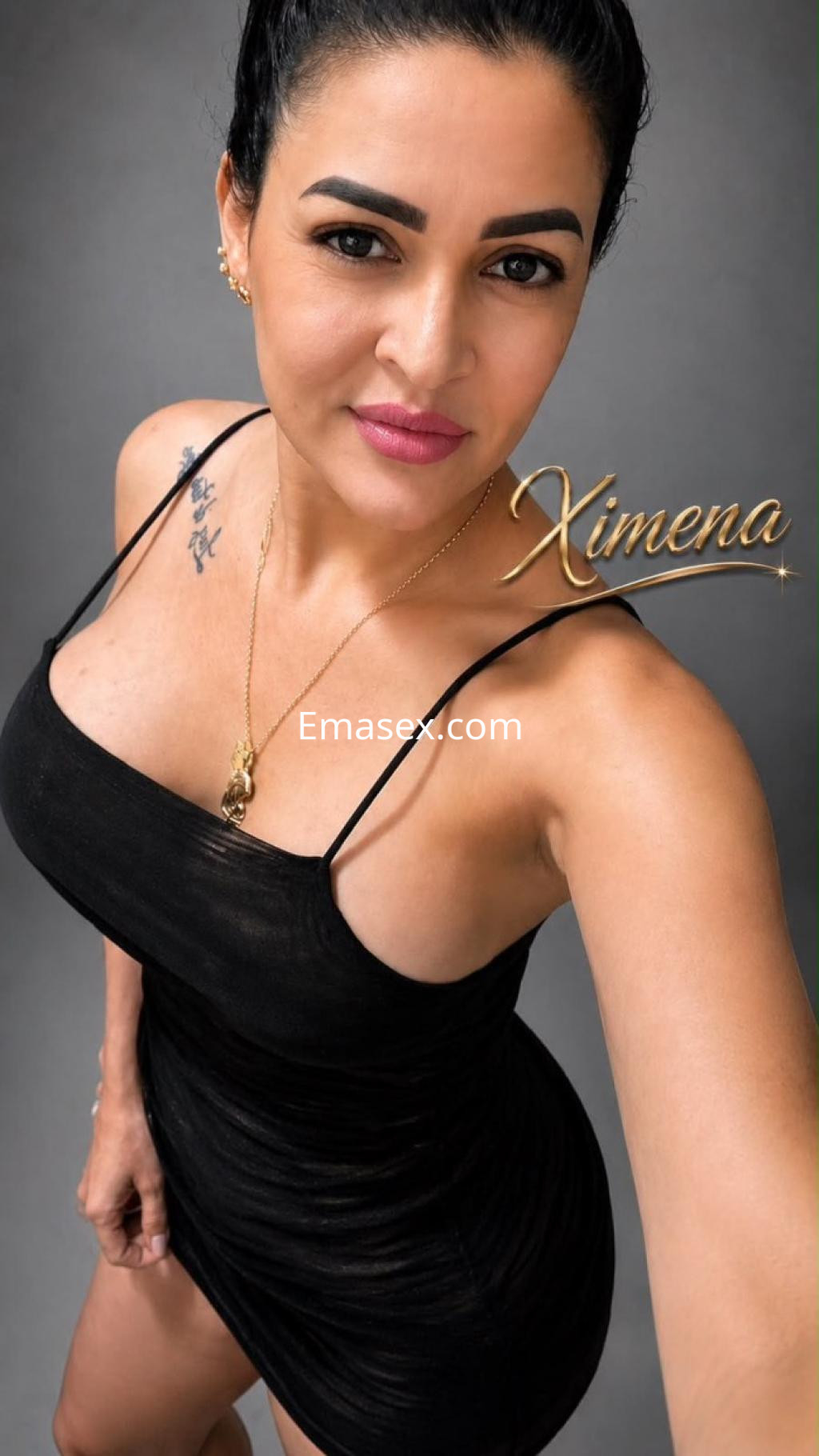 Escort en Huelva con un vestido negro ajustado, cabello recogido, posando con una sonrisa y tatuajes visibles.