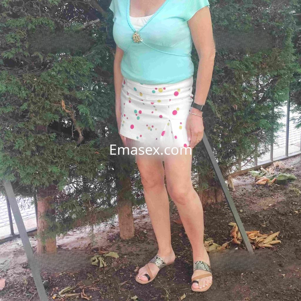 Puta en León, con blusa celeste y falda corta blanca con puntos de colores, de pie frente a un arbusto.