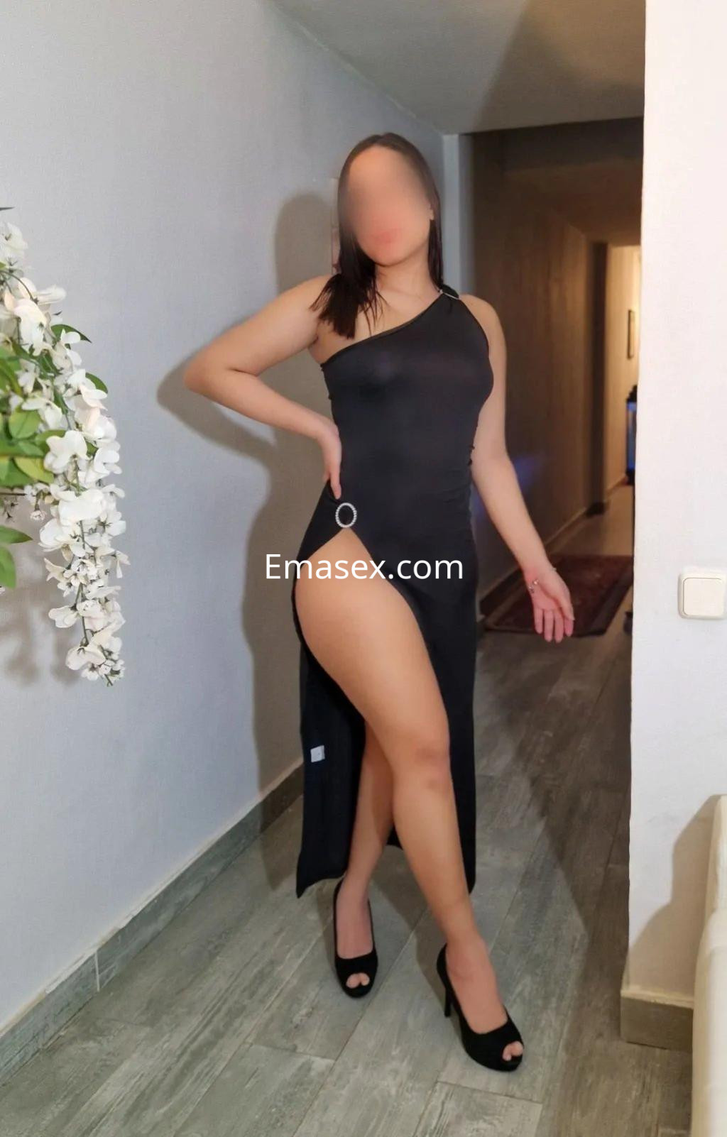 Escort en Madrid, luciendo un vestido negro de una sola manga y tacones altos, posando con una pierna al frente.