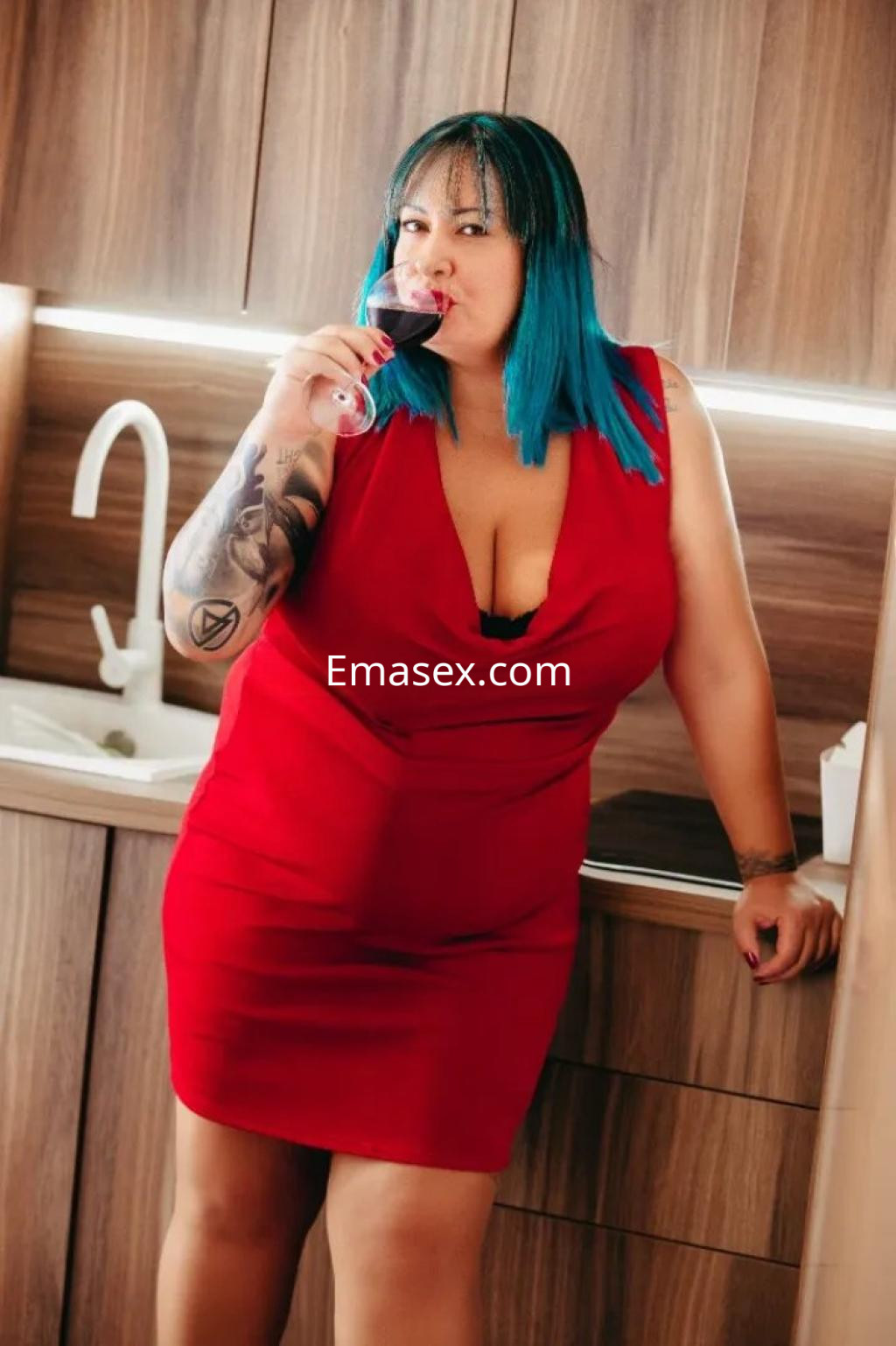 Escort en Sevilla con cabello azul y vestido rojo, posando en una cocina mientras sostiene una copa de vino.
