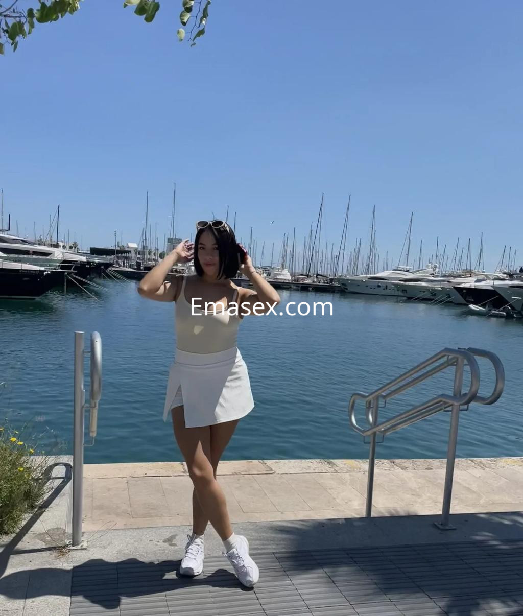 Escort en Alicante, posando frente al mar con un vestido blanco y blusa amarilla, rodeada de yates.