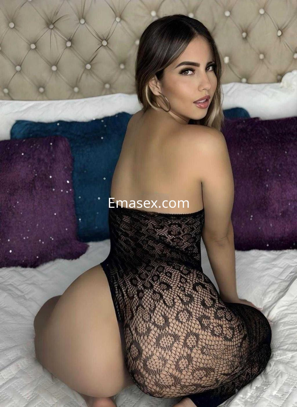 Escort en Barcelona, posando de espaldas sobre una cama con sábanas claras, luciendo un vestido de encaje negro.