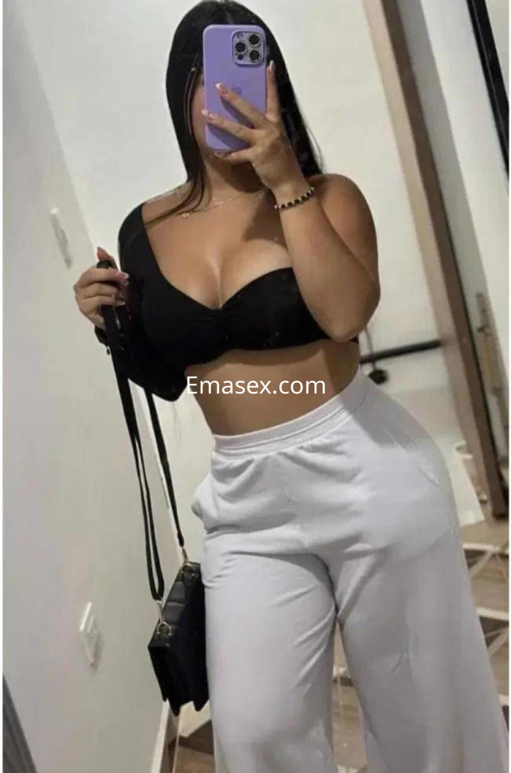 Escort en Burgos, luciendo un top negro y pantalones blancos, posando frente a un espejo con una bolsa negra.