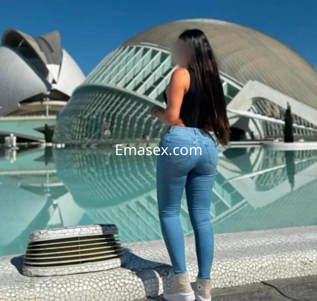 Escort de Sevilla posa de espaldas frente a una moderna estructura arquitectónica, luciendo jeans ajustados y camiseta.