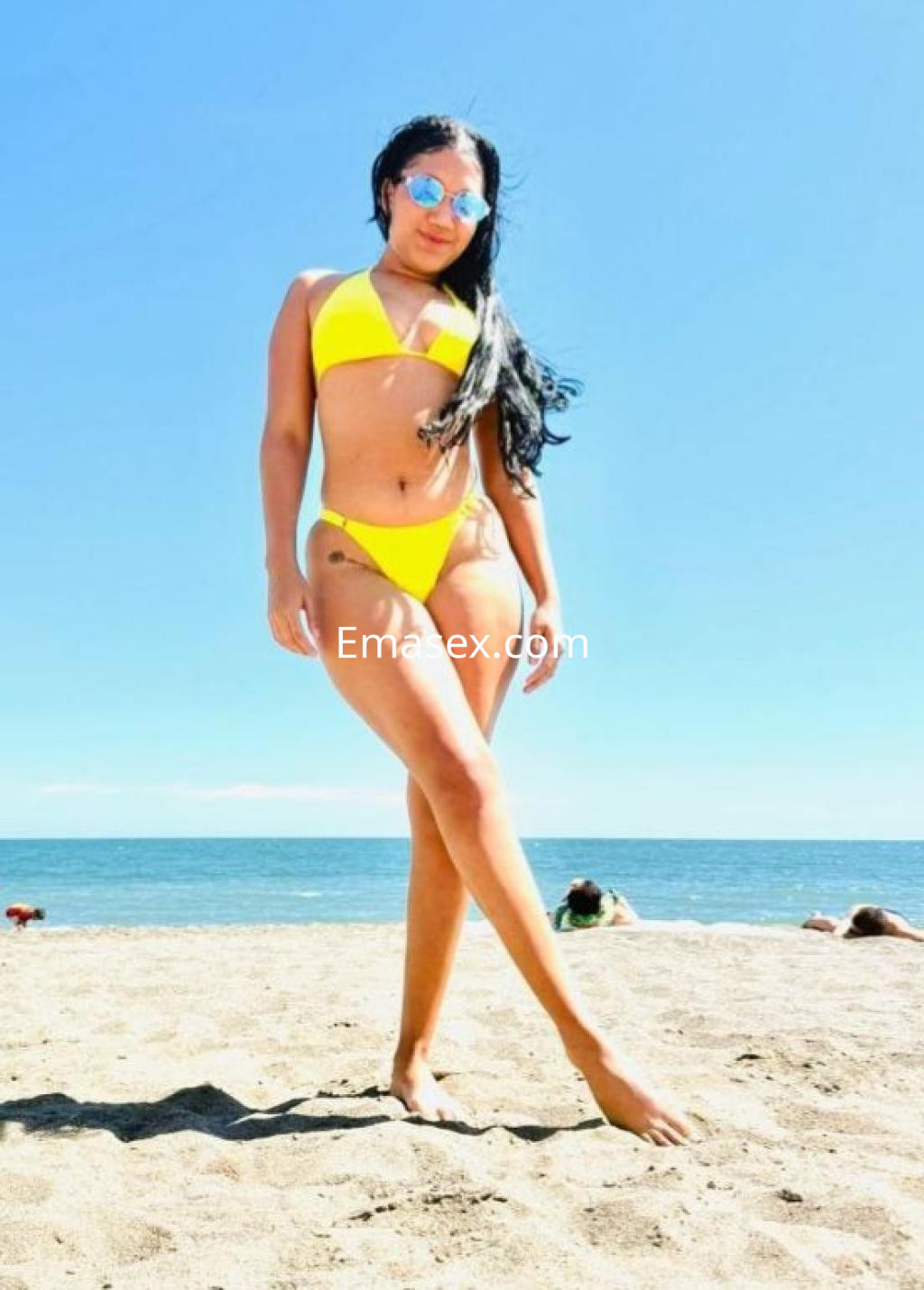 Puta posando en la playa de Málaga con bikini amarillo y gafas de sol, luciendo bronceada y sonriente.