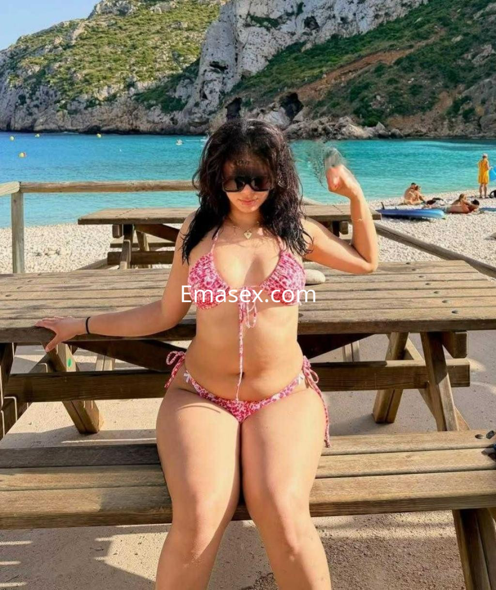 Escort en Madrid, sentada en una mesa de madera en la playa, con bikini rosa y gafas de sol, disfrutando del sol.