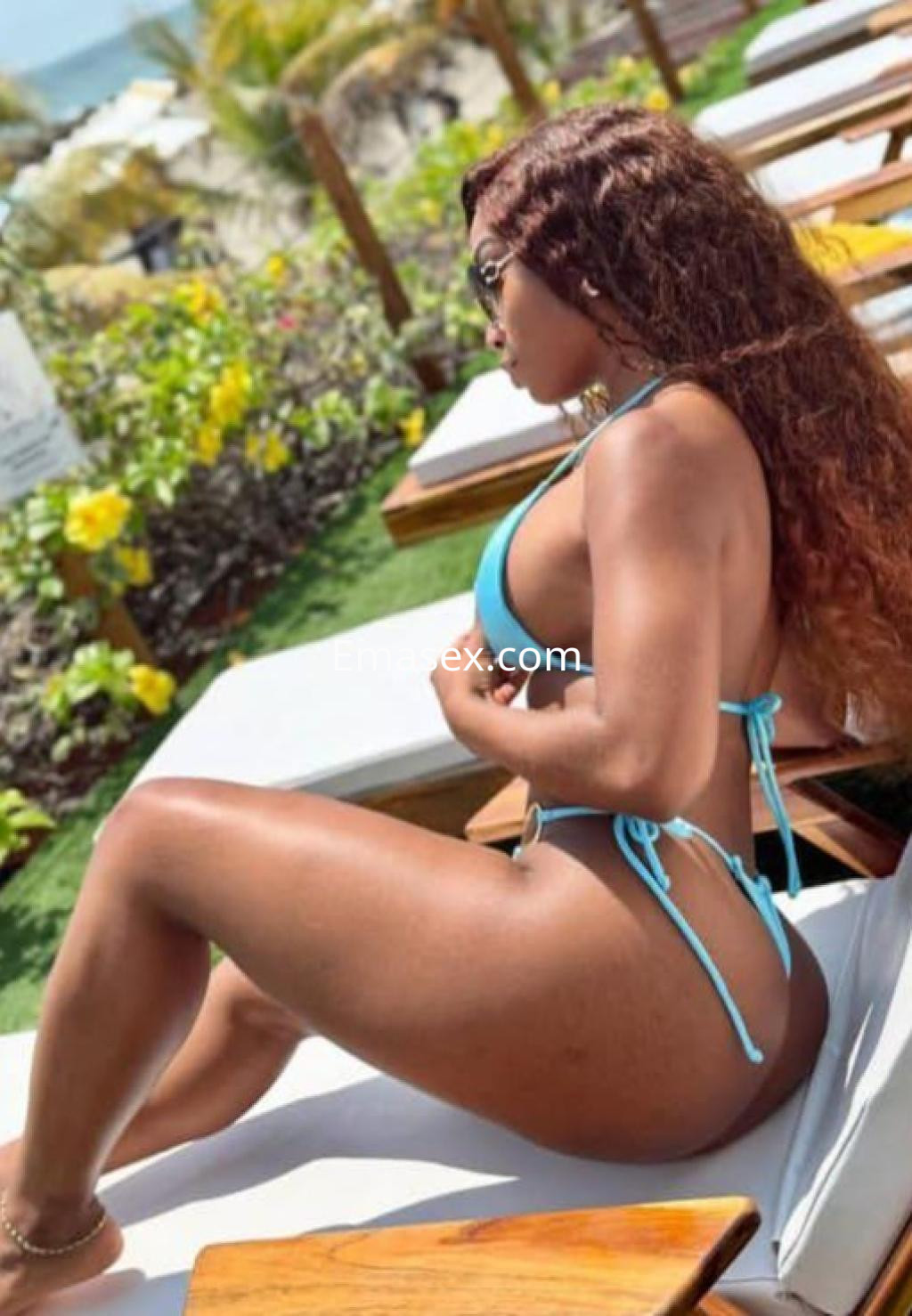 Escort en Girona, con cabello rizado y bronceado, posando en bikini azul junto a una piscina y flores.