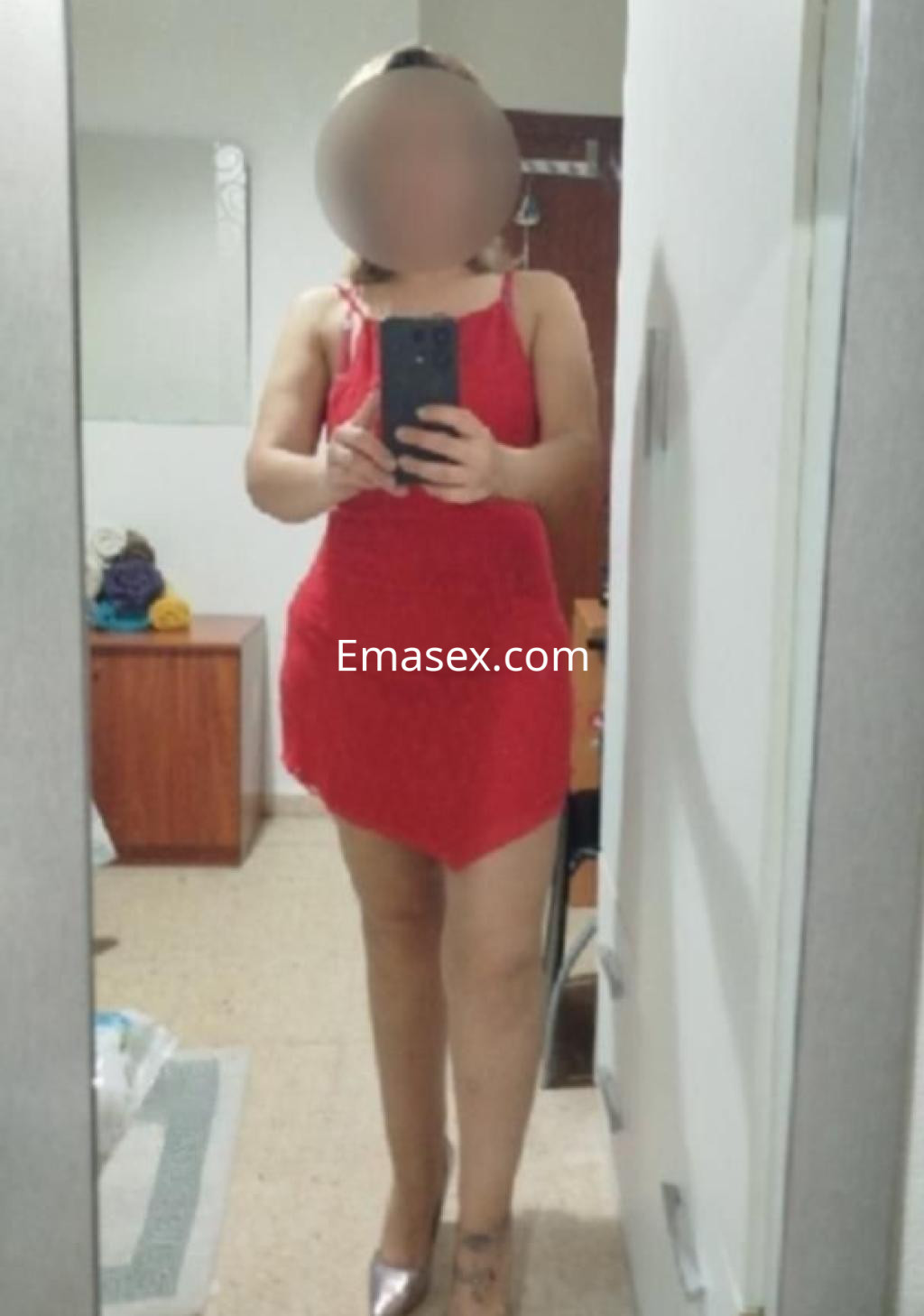 Puta en Huelva, posando frente al espejo con un vestido rojo y tacones, mostrando su figura.