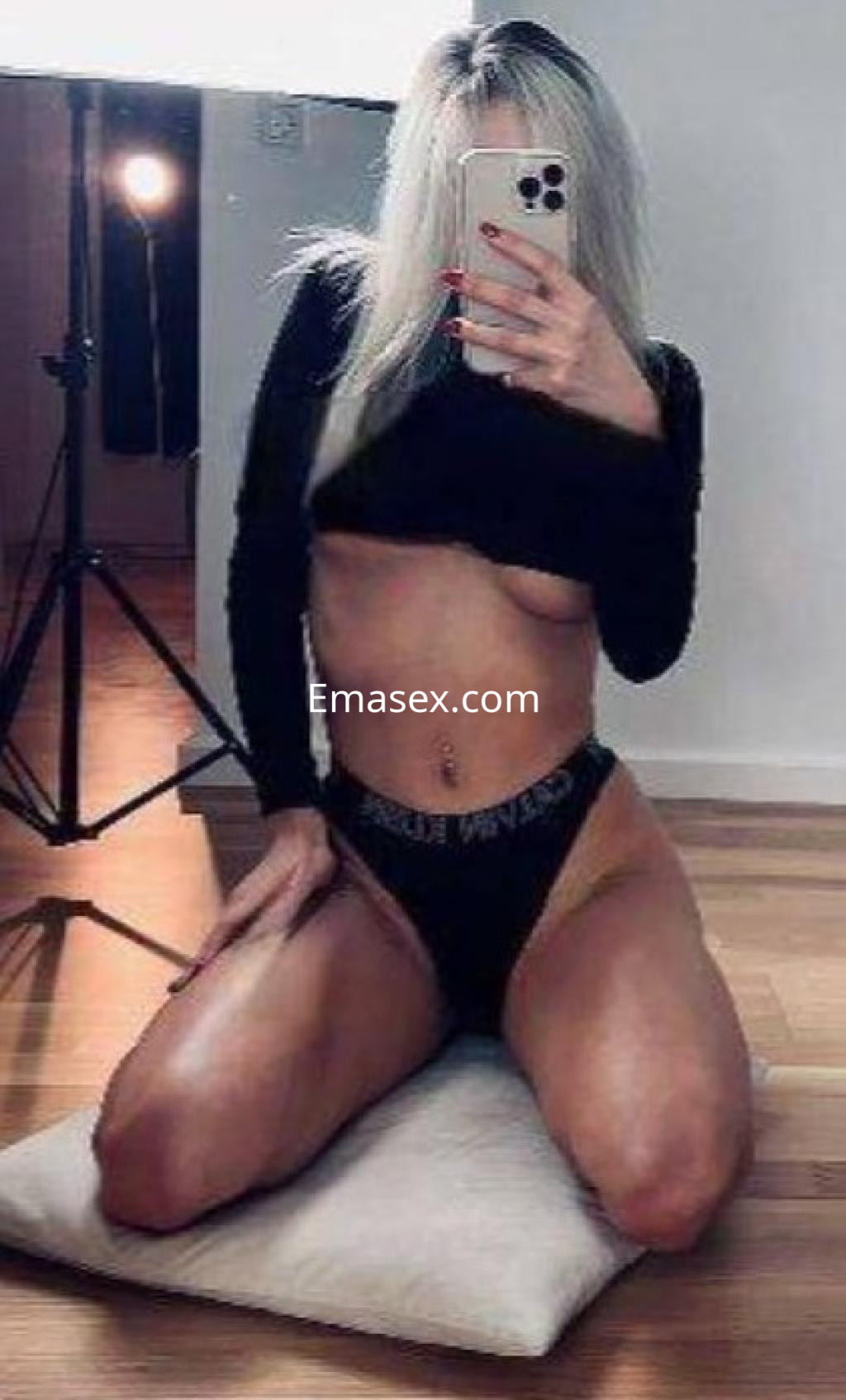Escort en Córdoba, posando de rodillas en un ambiente minimalista, con cabello rubio y atuendo negro.