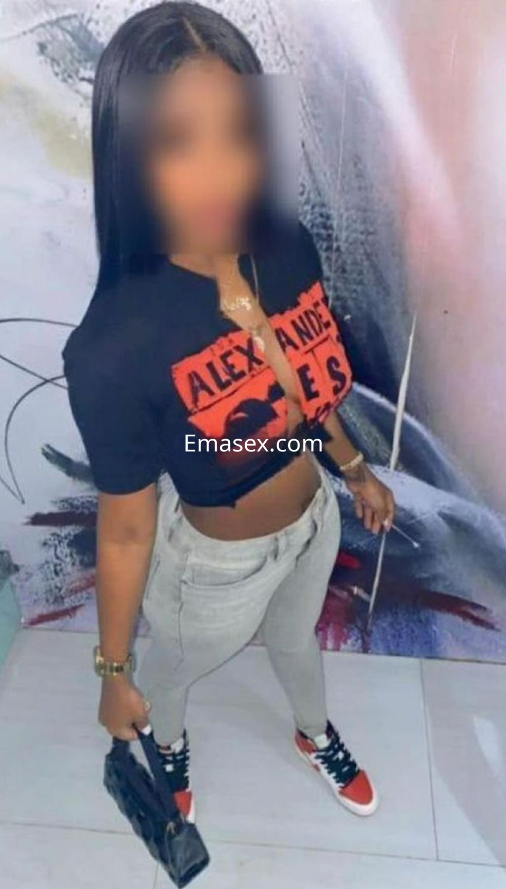Puta en Barcelona viste una camiseta corta negra y pantalones grises, sosteniendo una bolsa en un fondo artístico.