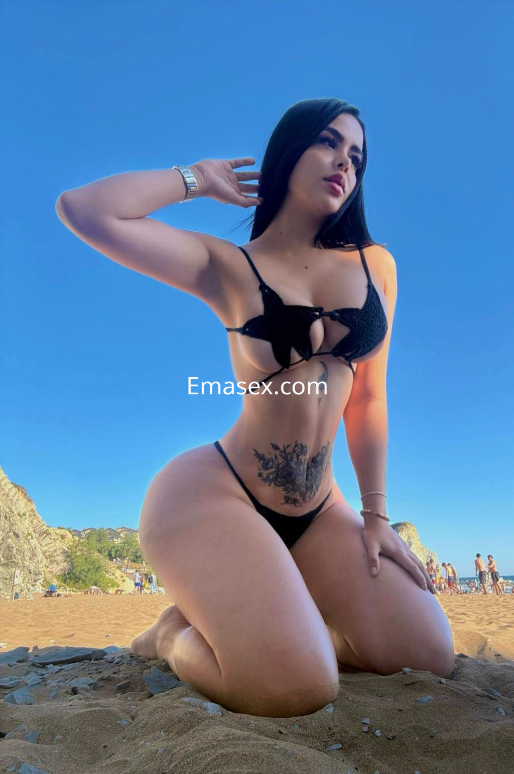 Puta en Bilbao posando de rodillas en la playa, luciendo un bikini negro y mostrando un tatuaje en el abdomen.