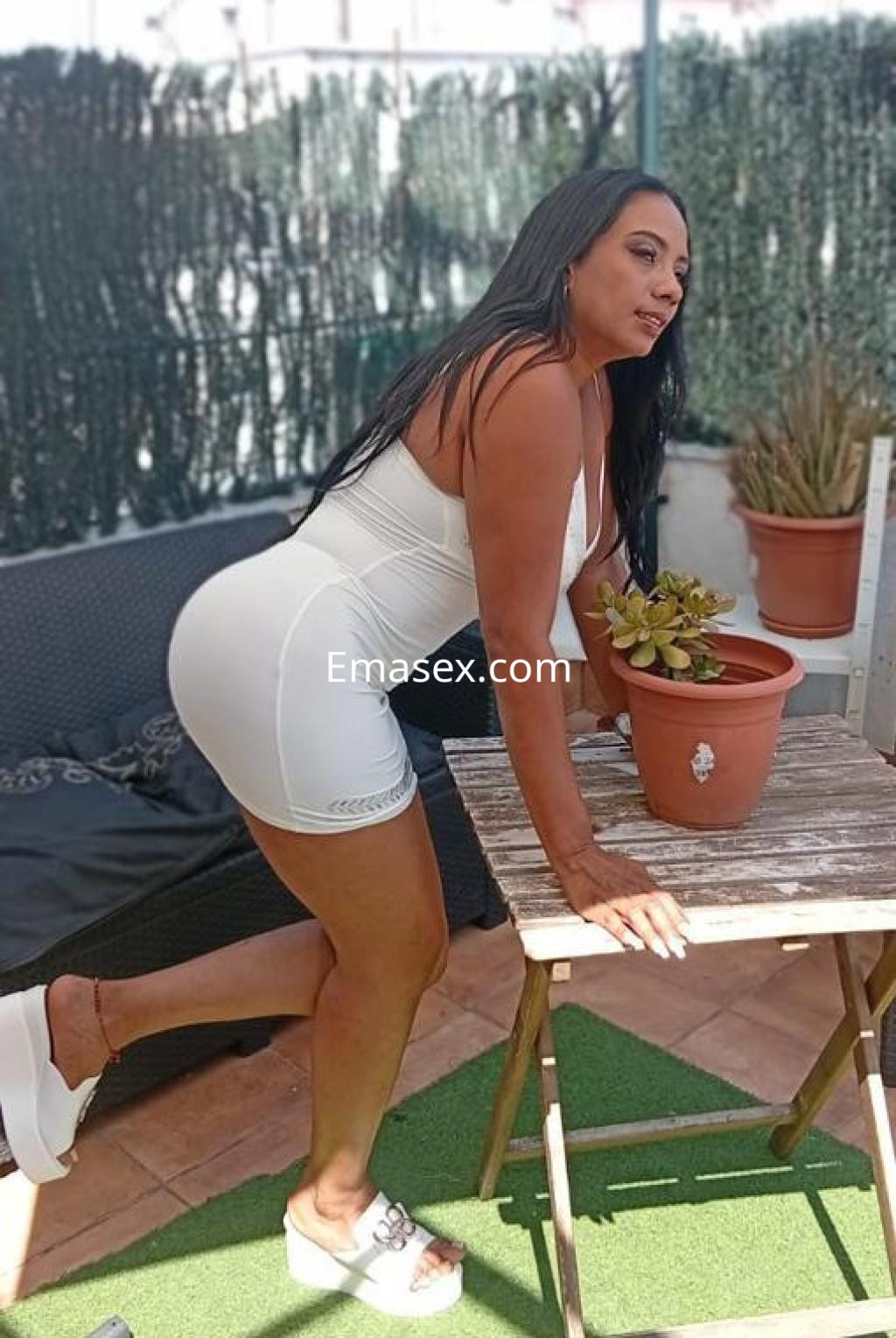 Escort en Barcelona, de cabello largo y oscuro, viste un conjunto blanco ajustado mientras se apoya en una mesa.