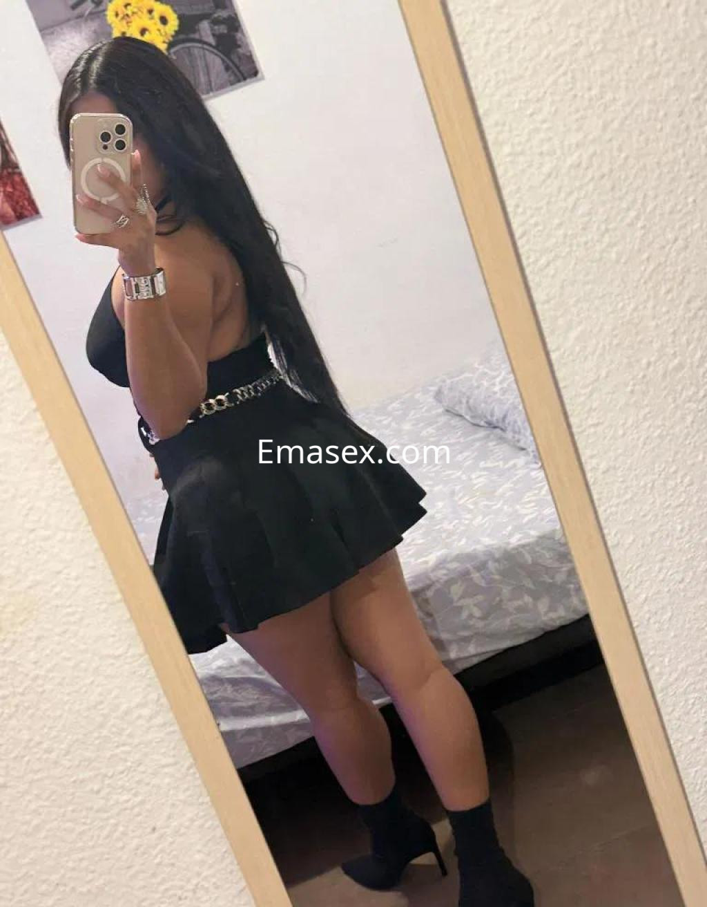 La puta en Huelva se muestra de espaldas en un espejo, luciendo una falda corta y blusa negra, con cabello largo.