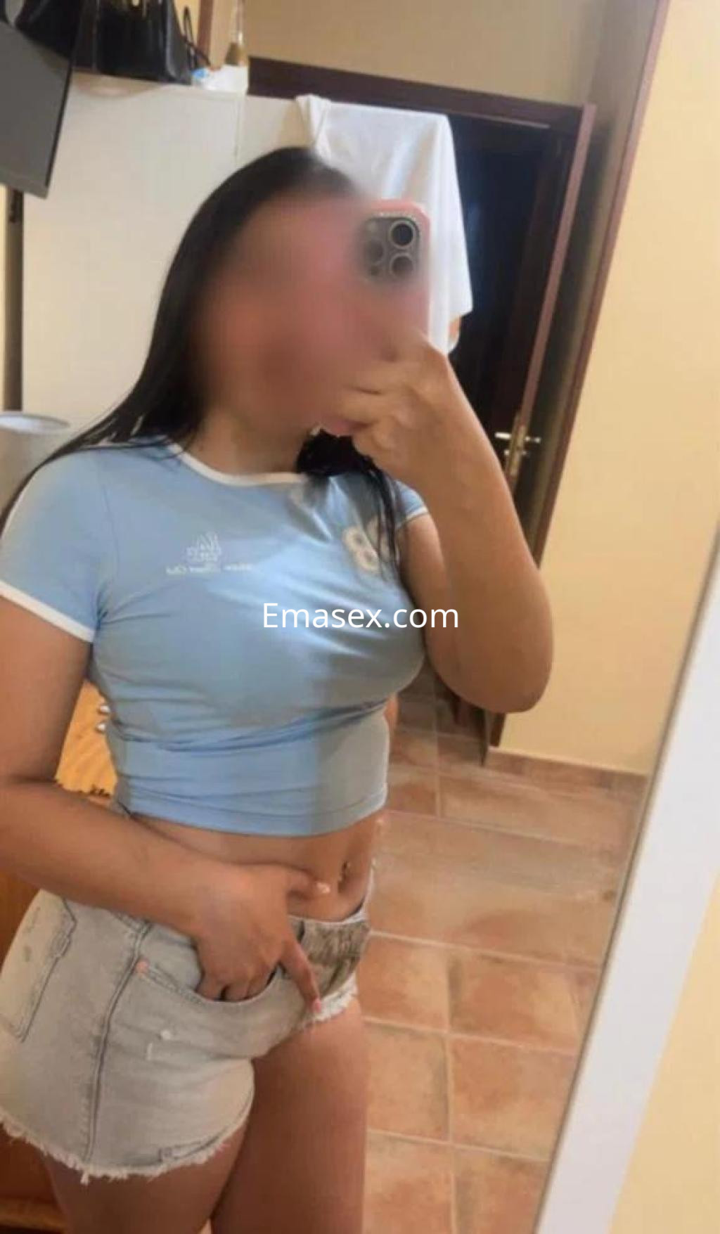 Puta en Murcia con camiseta azul y shorts cortos, posando frente a un espejo en un entorno interior.