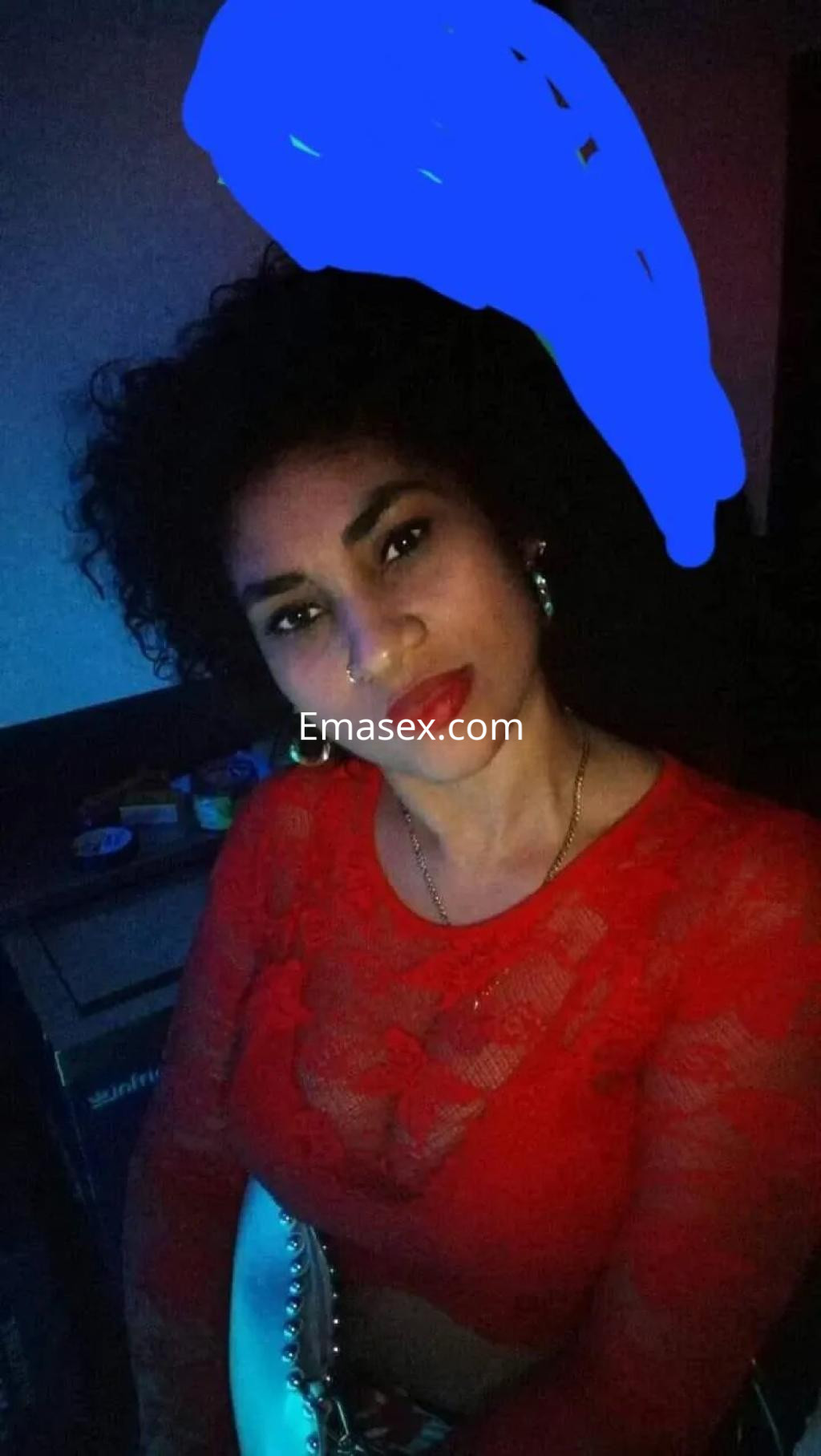 Puta con cabello rizado y oscuro, vestida con top rojo de encaje, posando en un ambiente tenue en Oviedo.