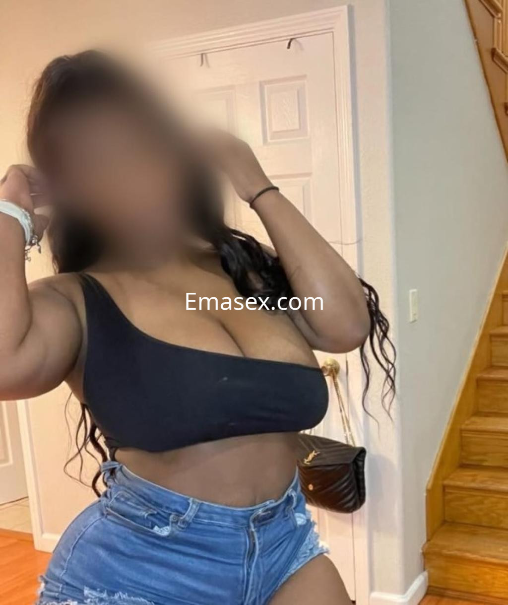 Escort en Oviedo con cabello largo y ondulado, vistiendo un top negro y shorts de mezclilla, posando en un interior.