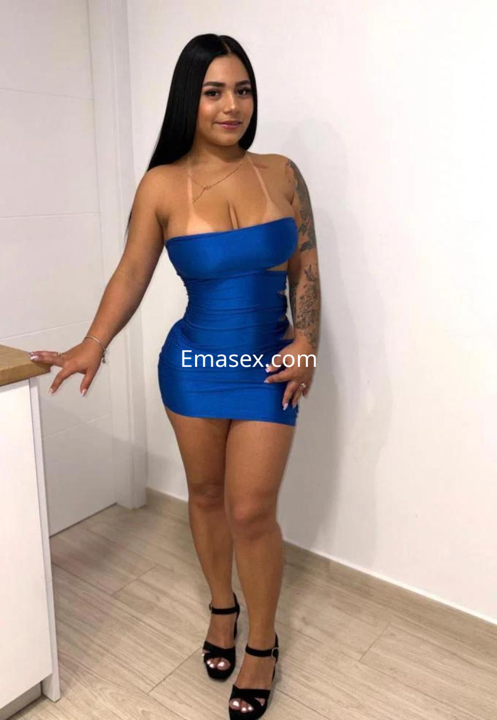 Escort en Las Palmas con vestido azul ajustado, tacones negros y cabello lacio, posando junto a una pared blanca.