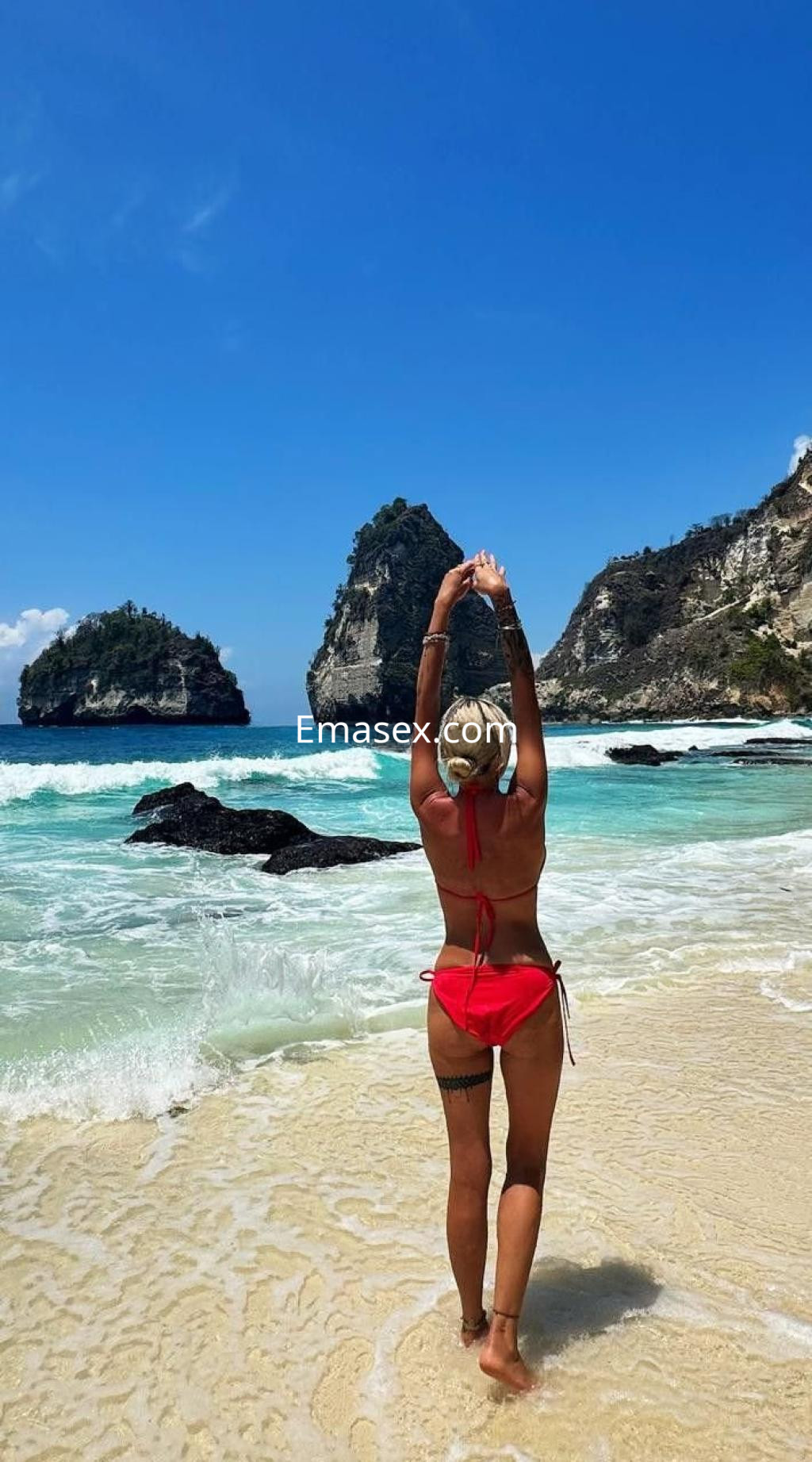 Escort en Valencia con un bikini rojo, de pie en la playa con los brazos levantados, mirando al mar.