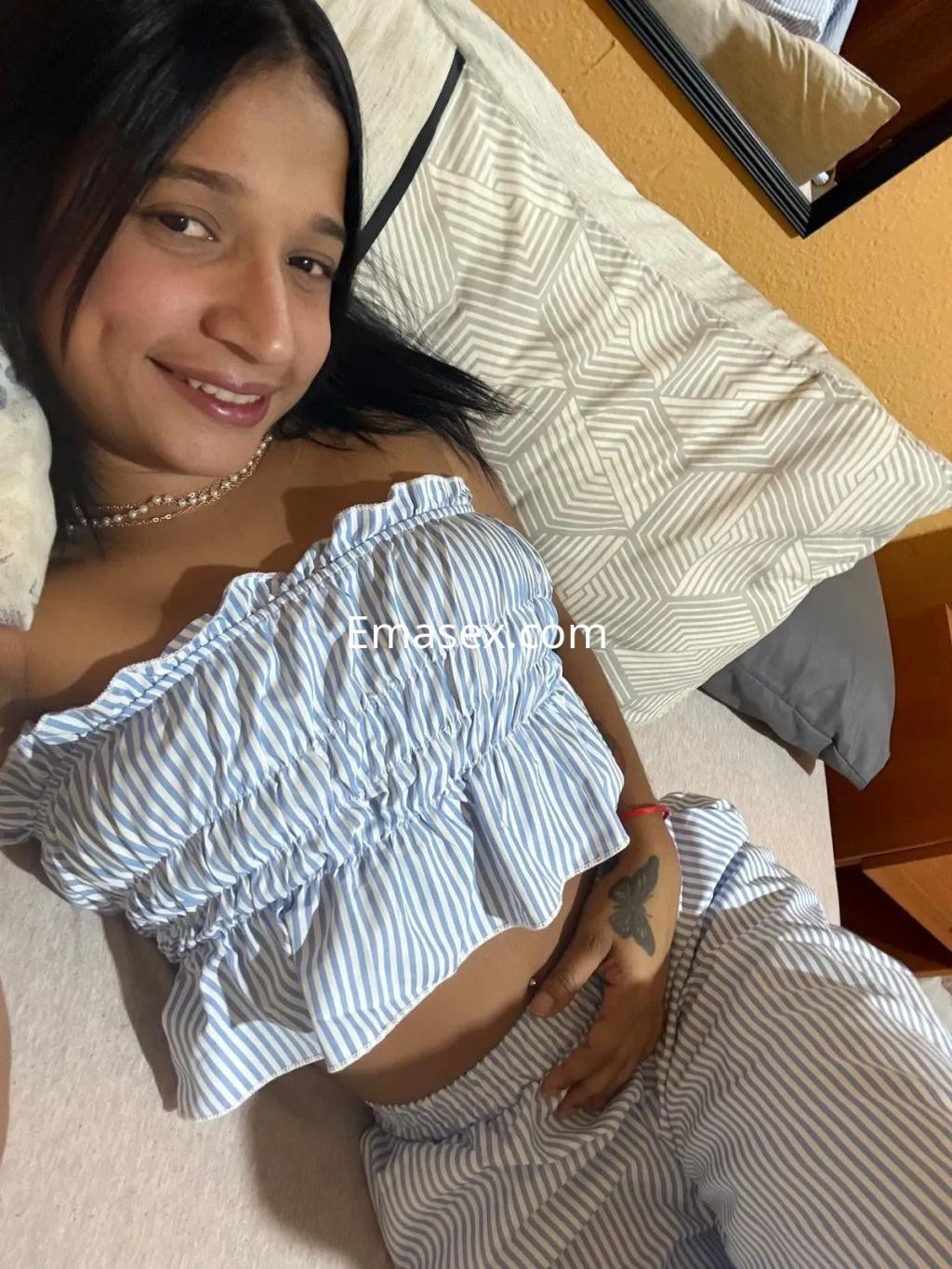 Escort en Zaragoza, posando sonriente sobre una cama, lleva un conjunto de blusa y pantalones a rayas.