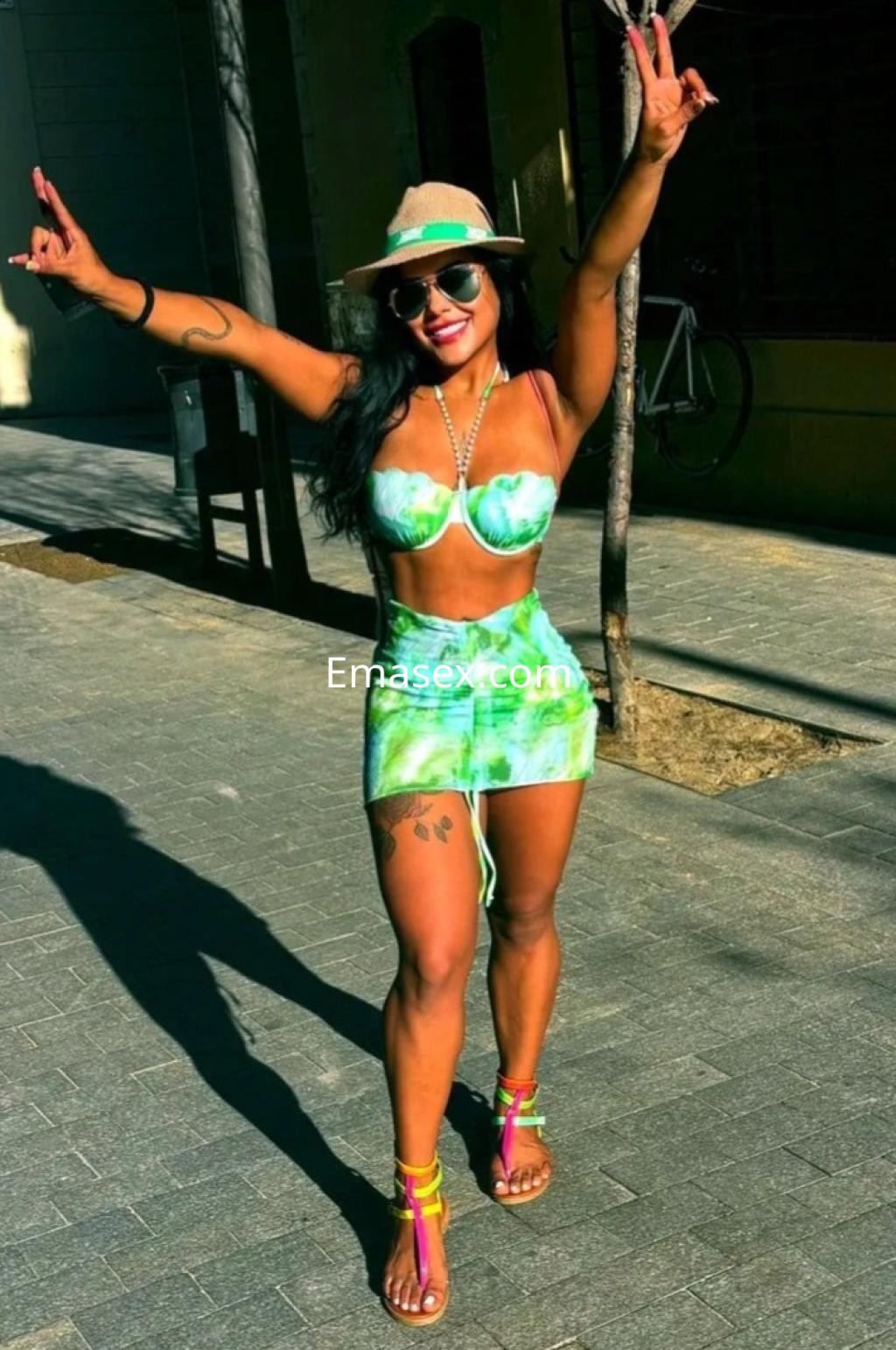 Puta sonriente en Granada, vistiendo un conjunto de bikini verde y una falda corta, con sombrero y gafas de sol.