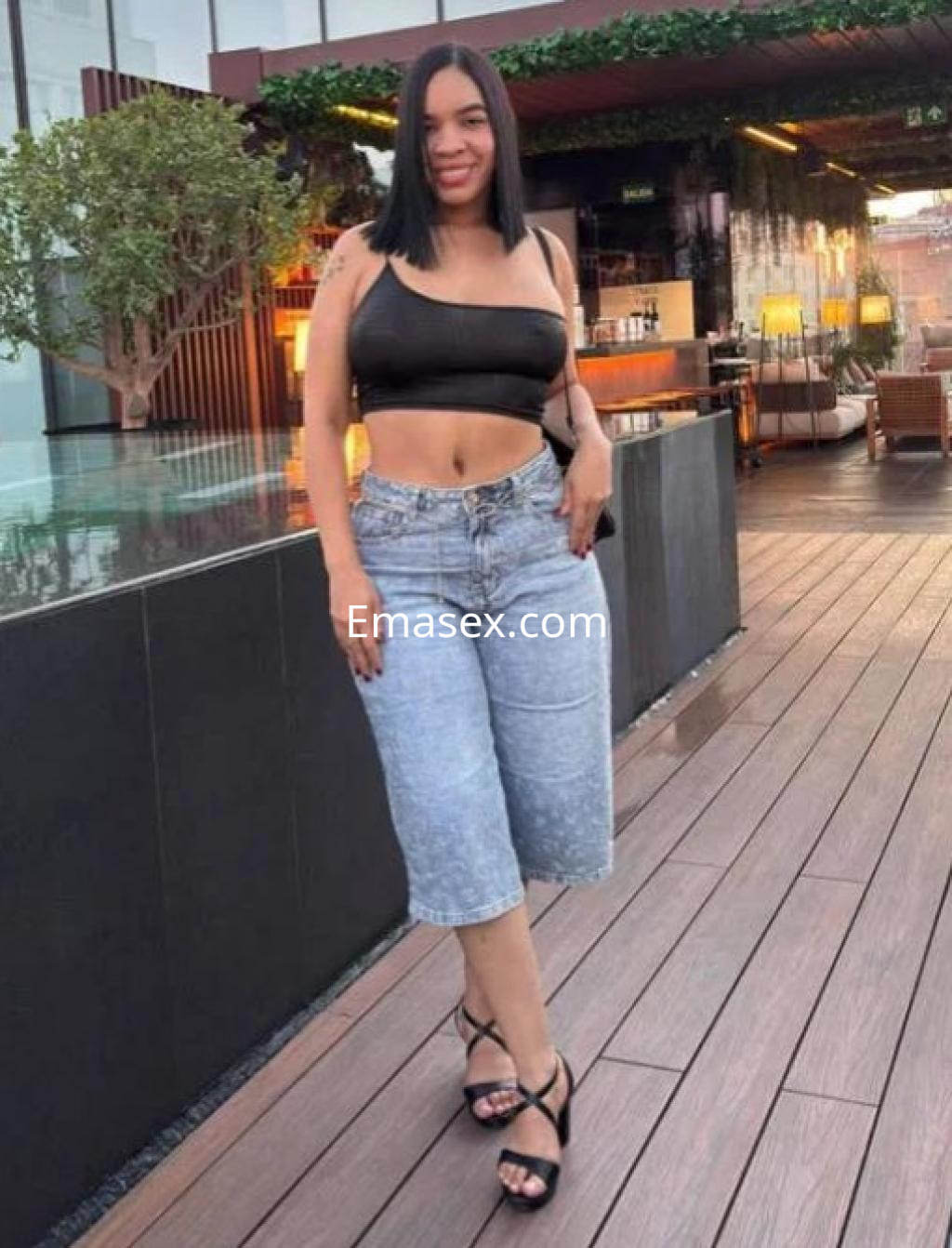 Escort en Sevilla vistiendo top negro y pantalones cortos de mezclilla, posando en un ambiente exterior elegante.