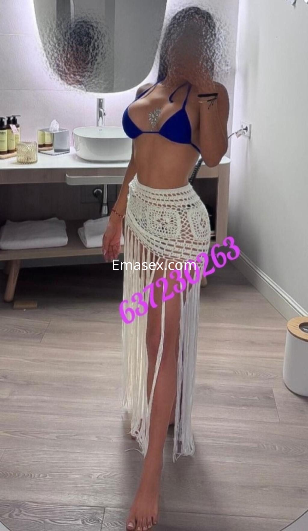 Puta en Lleida luce un bikini azul con una falda de hilo blanco, posando frente a un espejo en un baño moderno.