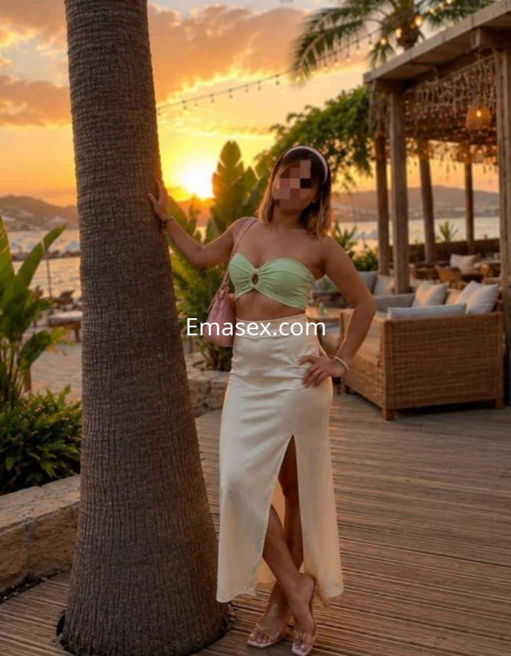 Escort en Madrid posando junto a una palmera al atardecer, con un top verde claro y una falda blanca.