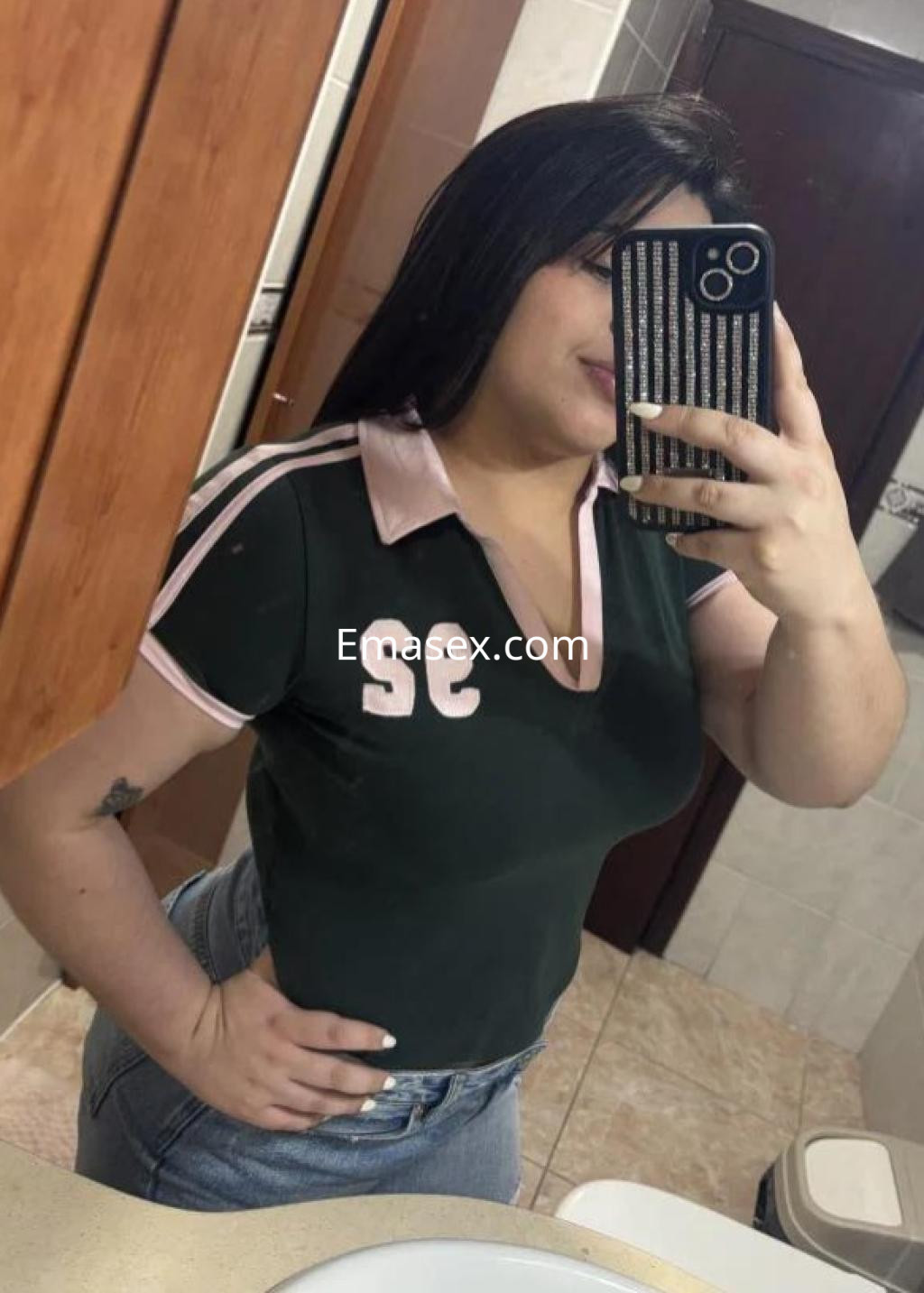 Escort en Las Palmas posando frente al espejo, lleva una camiseta verde con detalles rosas y jeans ajustados.