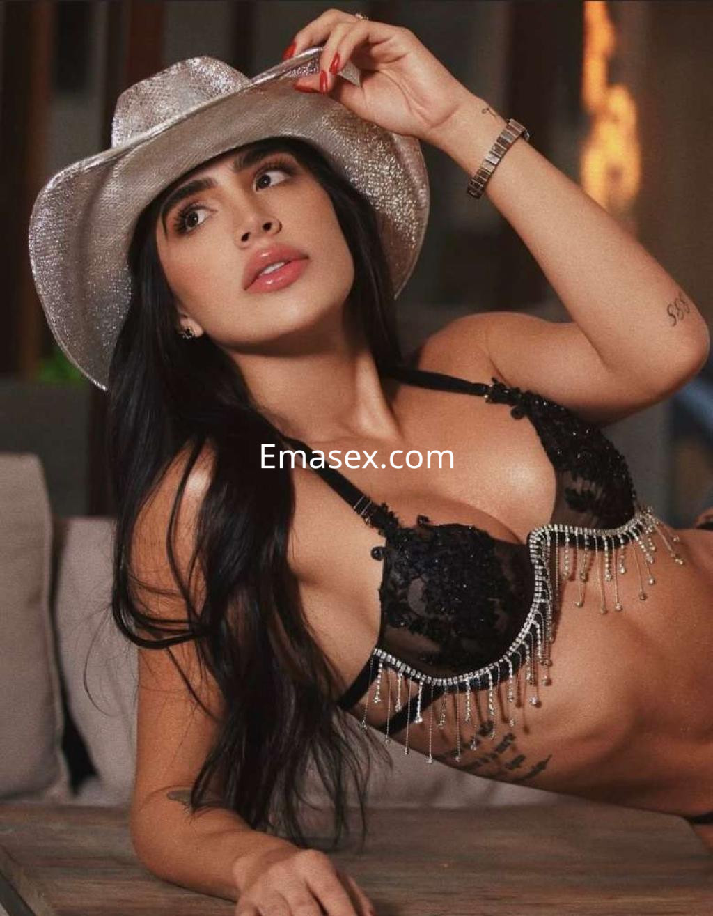 Puta en Ibiza recostada sobre un sofá, luciendo un sujetador negro con detalles brillantes y un sombrero plateado.