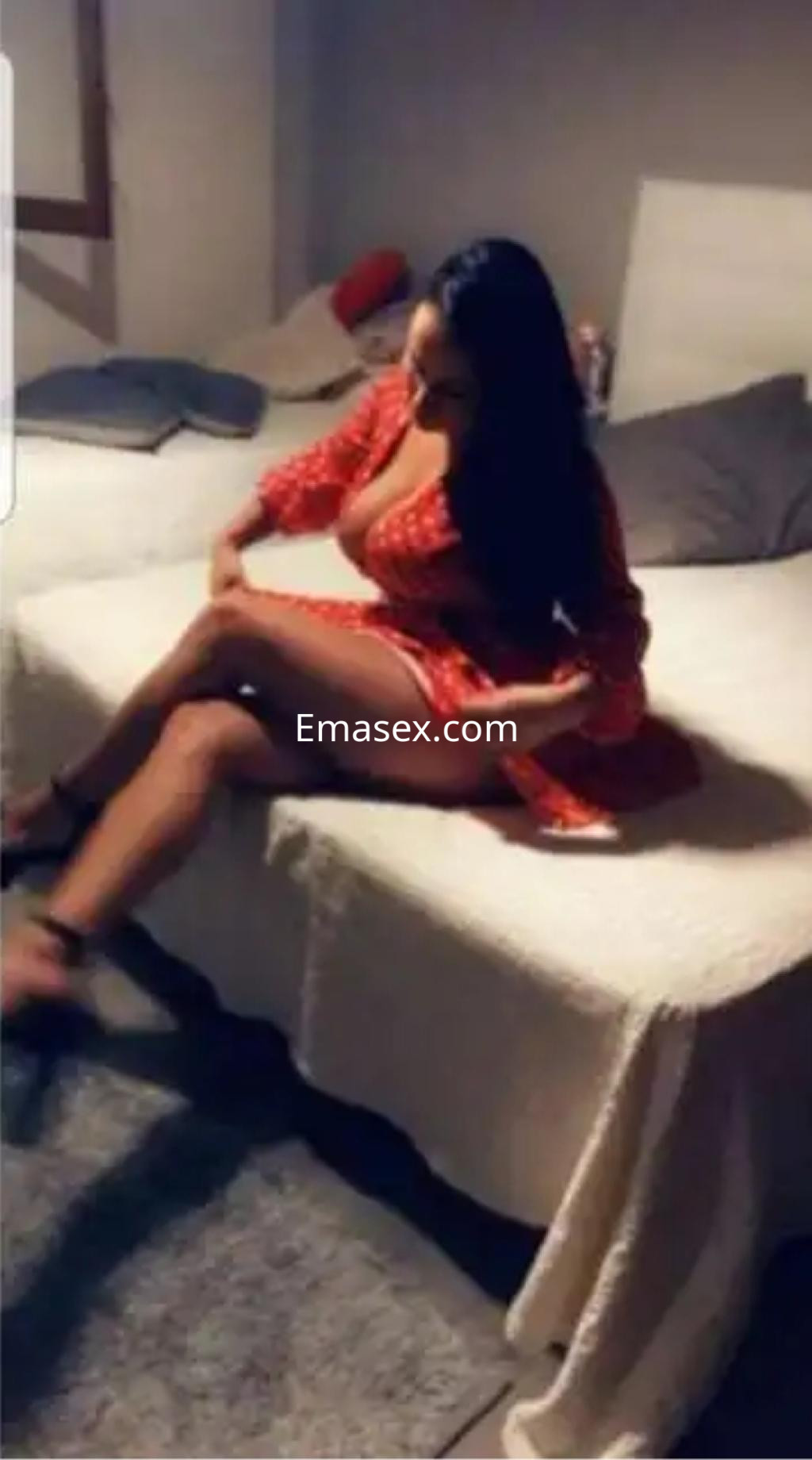 La escort en Murcia viste un vestido rojo mientras se sienta en la cama, mostrando una pose elegante.