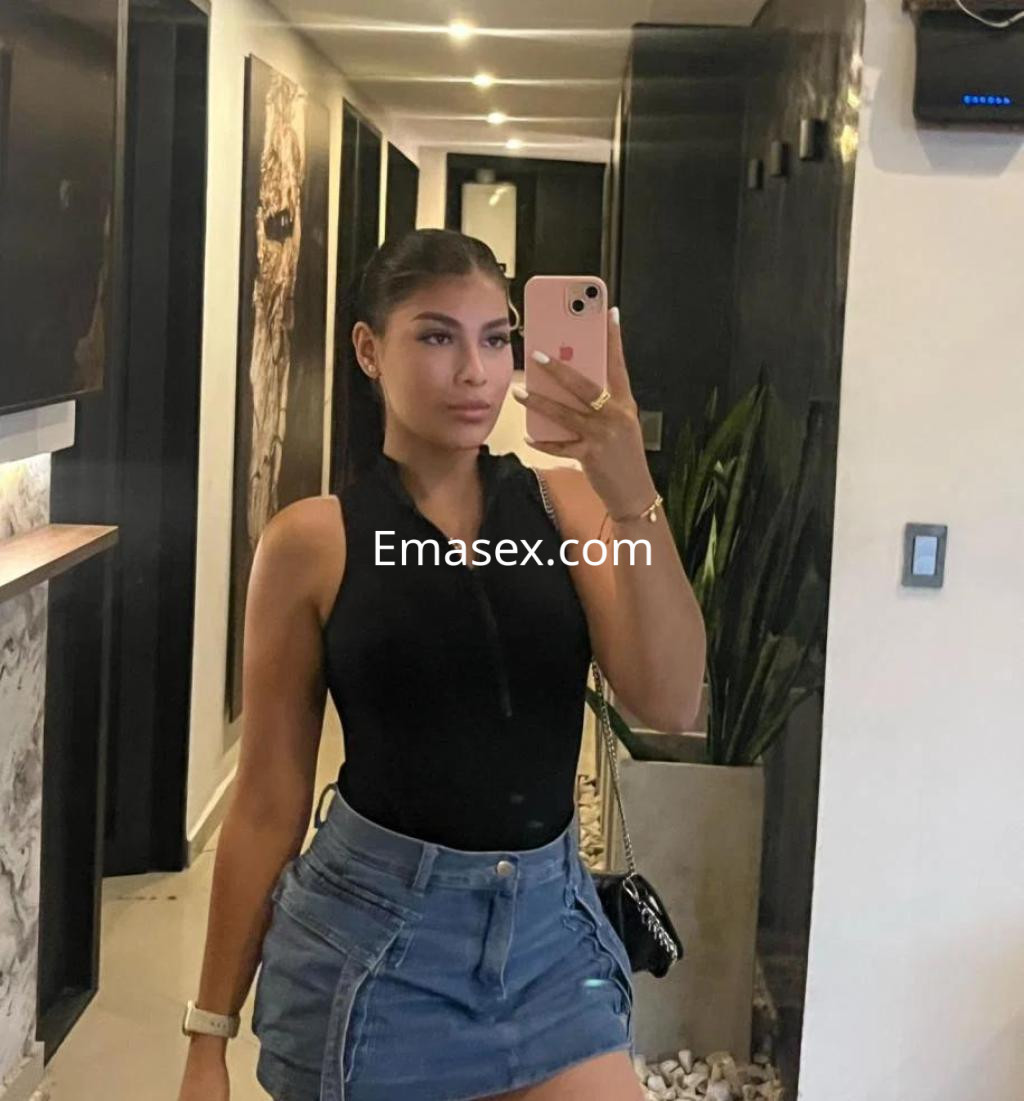 Escort en Alicante con cabello largo y lacio, lleva un body negro y una falda corta de mezclilla, sonriendo en un espejo.