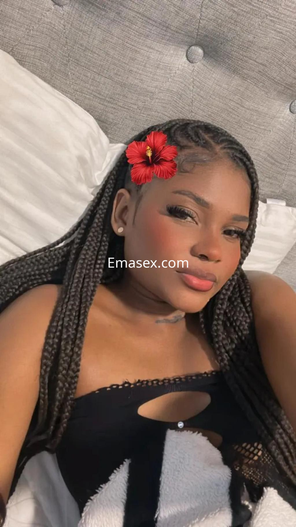 Puta en Alicante, con trenzas largas y un adorno floral en el cabello, posando en una cama con sábanas blancas.