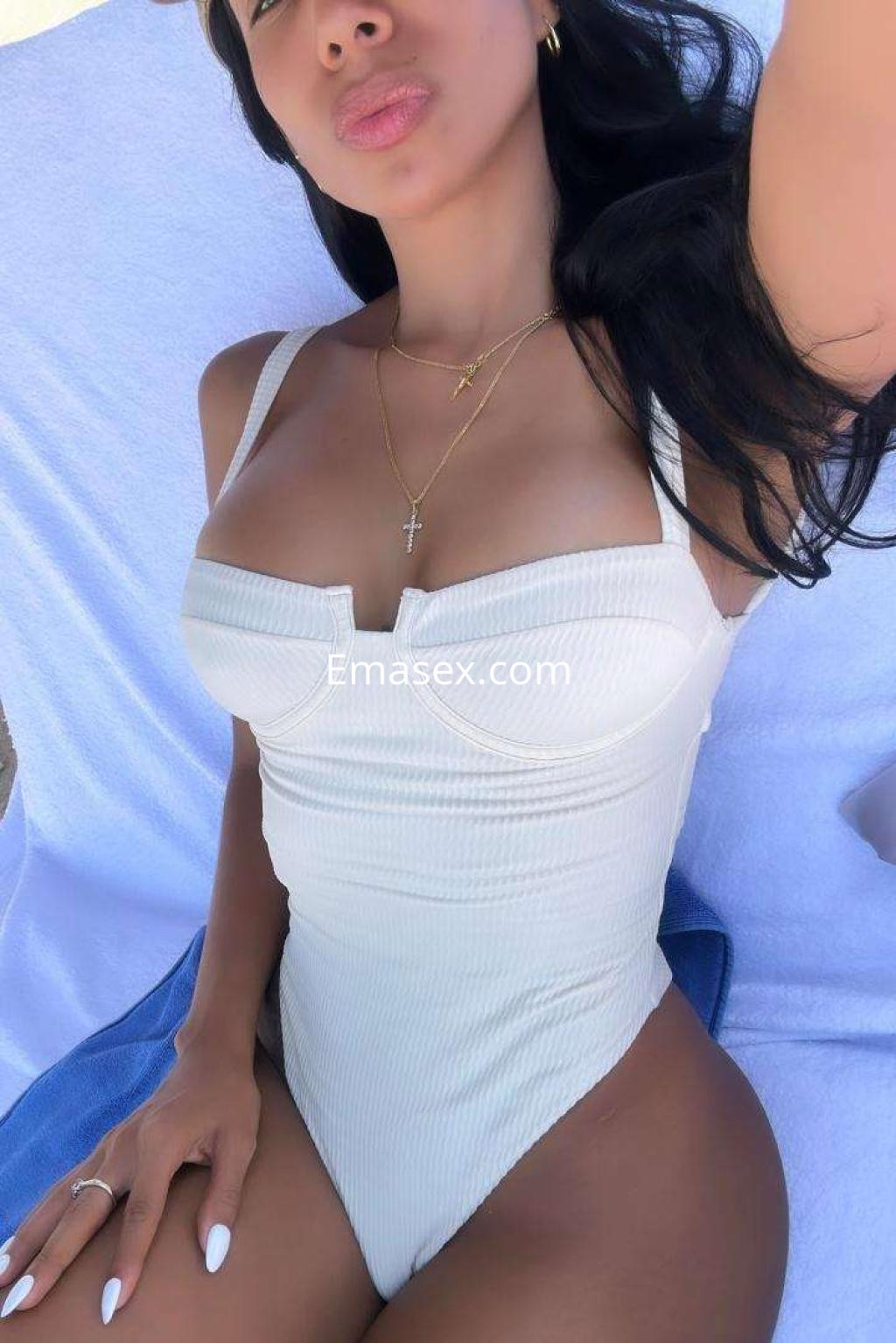 Escort en Ibiza, posando con un traje de baño blanco de tirantes, cabello largo y ondulado, con un fondo claro.