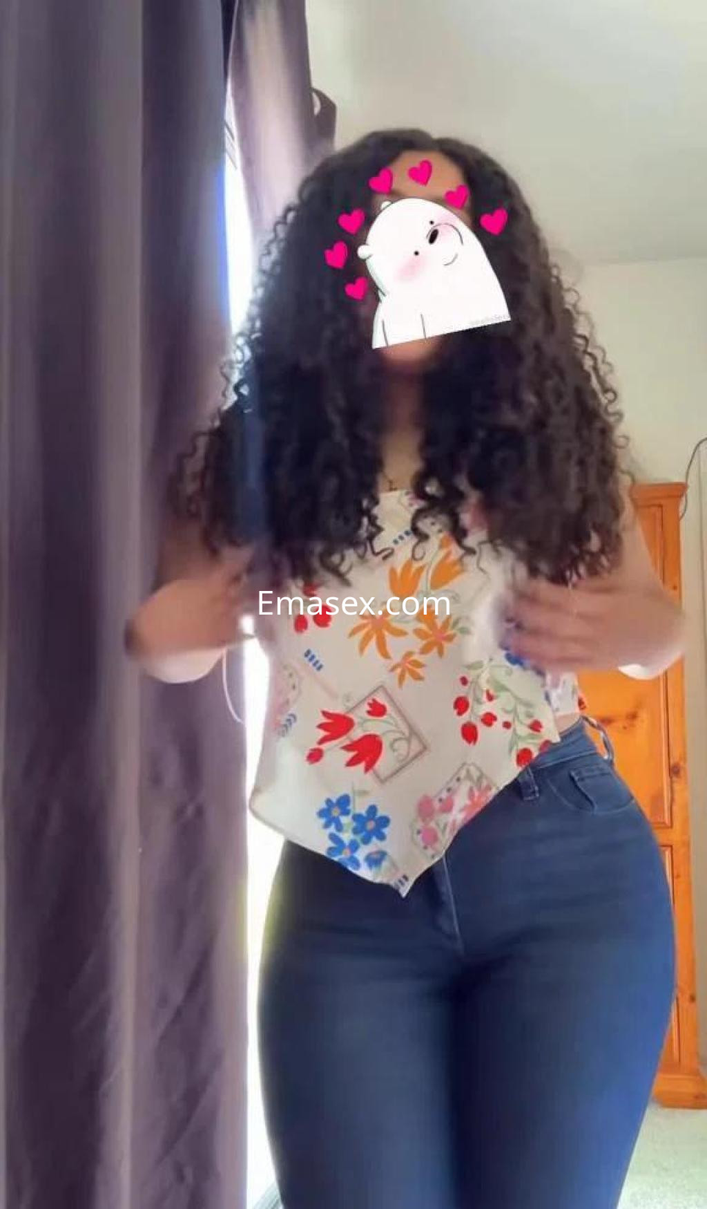 Puta con cabello rizado y largo, viste una blusa estampada y jeans ajustados, posando en una habitación iluminada en Cáceres.