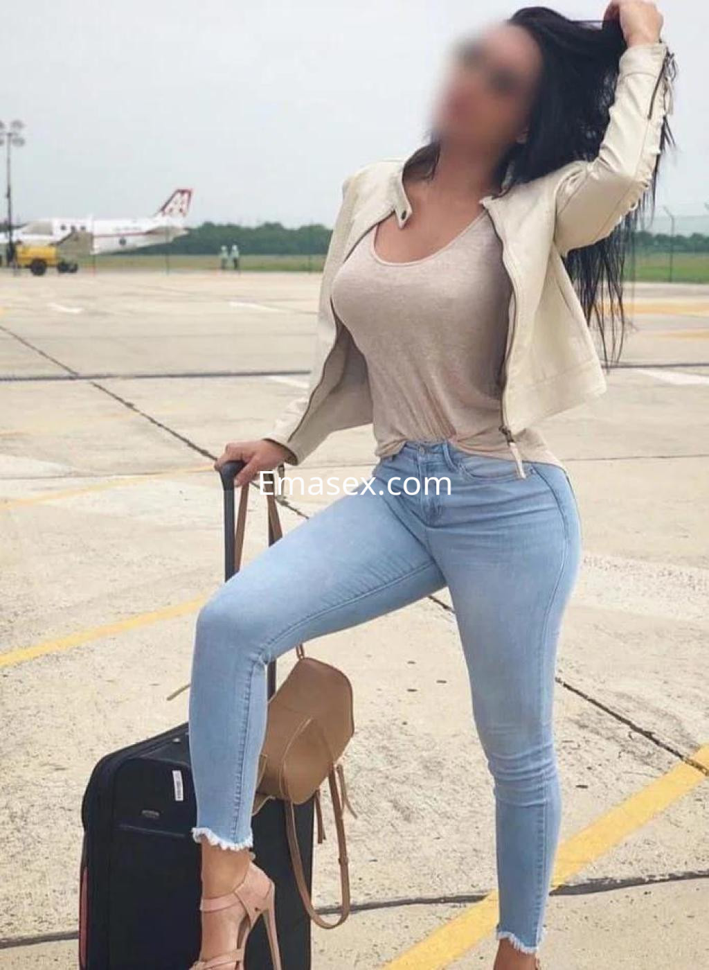 Escort en Córdoba posando en un aeropuerto, con blusa clara, jeans ajustados y una maleta de viaje.