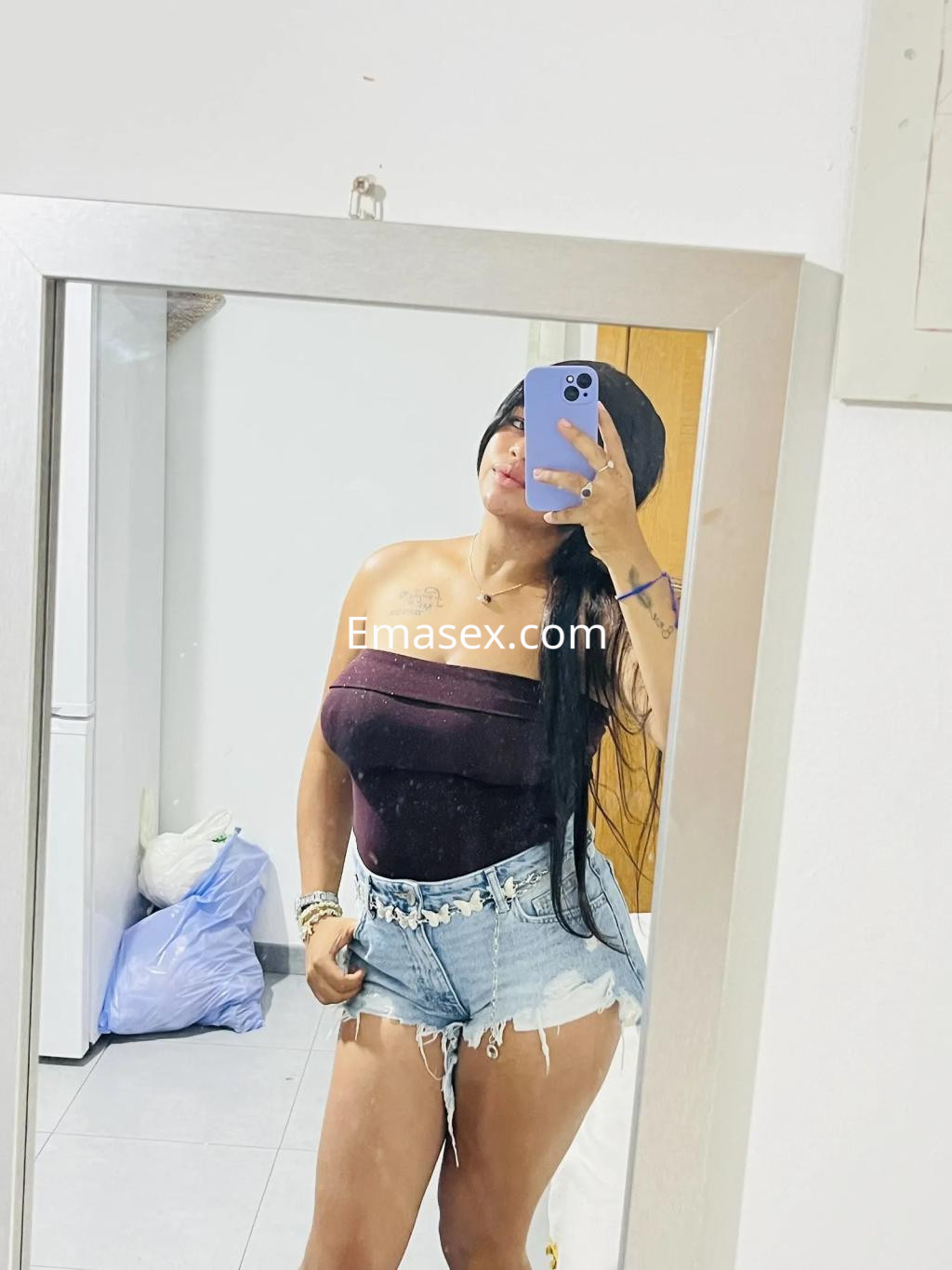 La puta en Córdoba posa frente al espejo con un top morado y shorts de mezclilla, mostrando su larga cabellera.