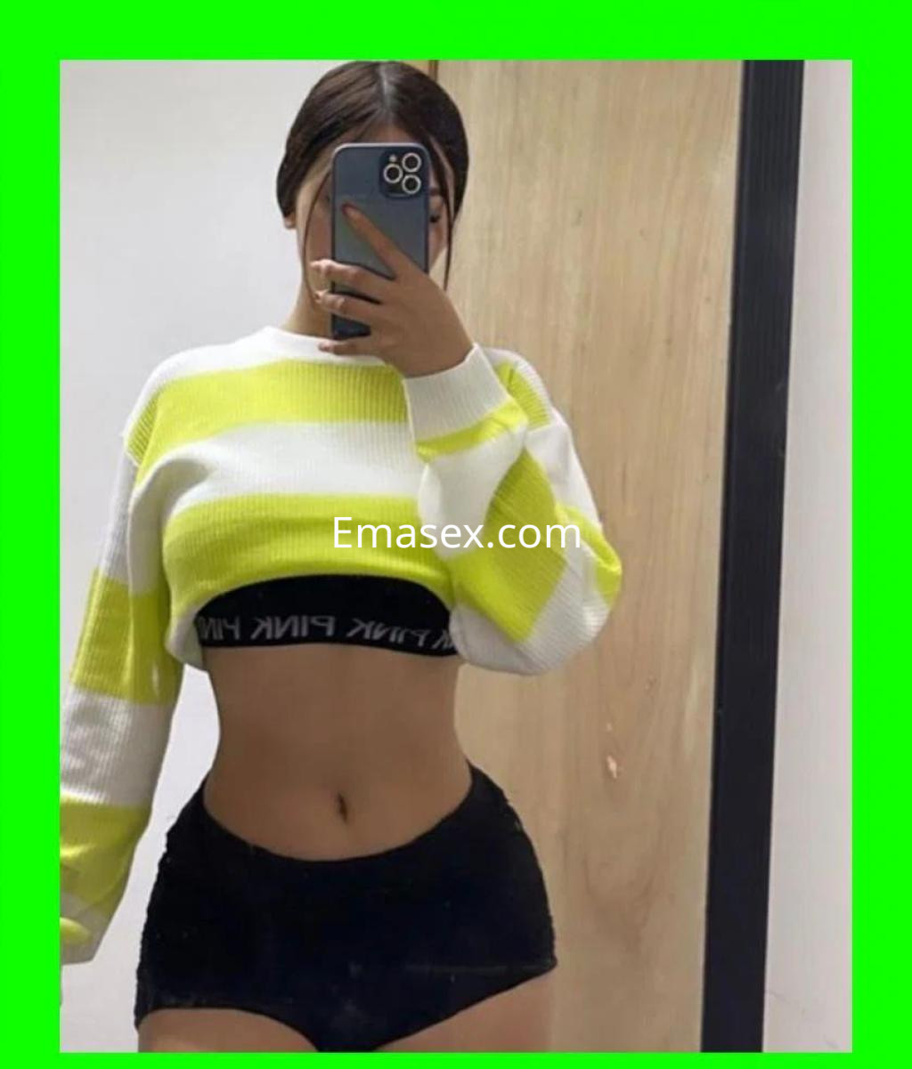 Escort en Coruña con cabello oscuro, viste un suéter blanco y amarillo con pantalones cortos negros, posando frente al espejo.
