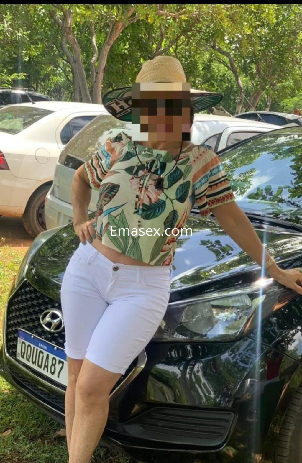 Escort en Coruña posando junto a un coche negro, vistiendo una camiseta de estampado floral y pantalones cortos blancos.