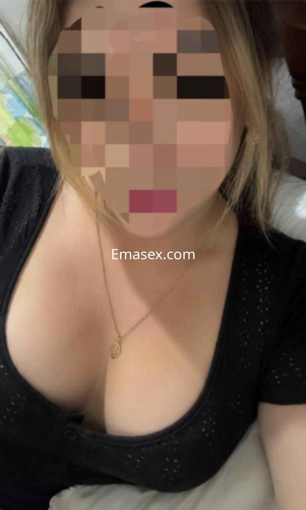 Puta en León con blusa negra ajustada que resalta su escote y un collar delicado, posando en la cama.