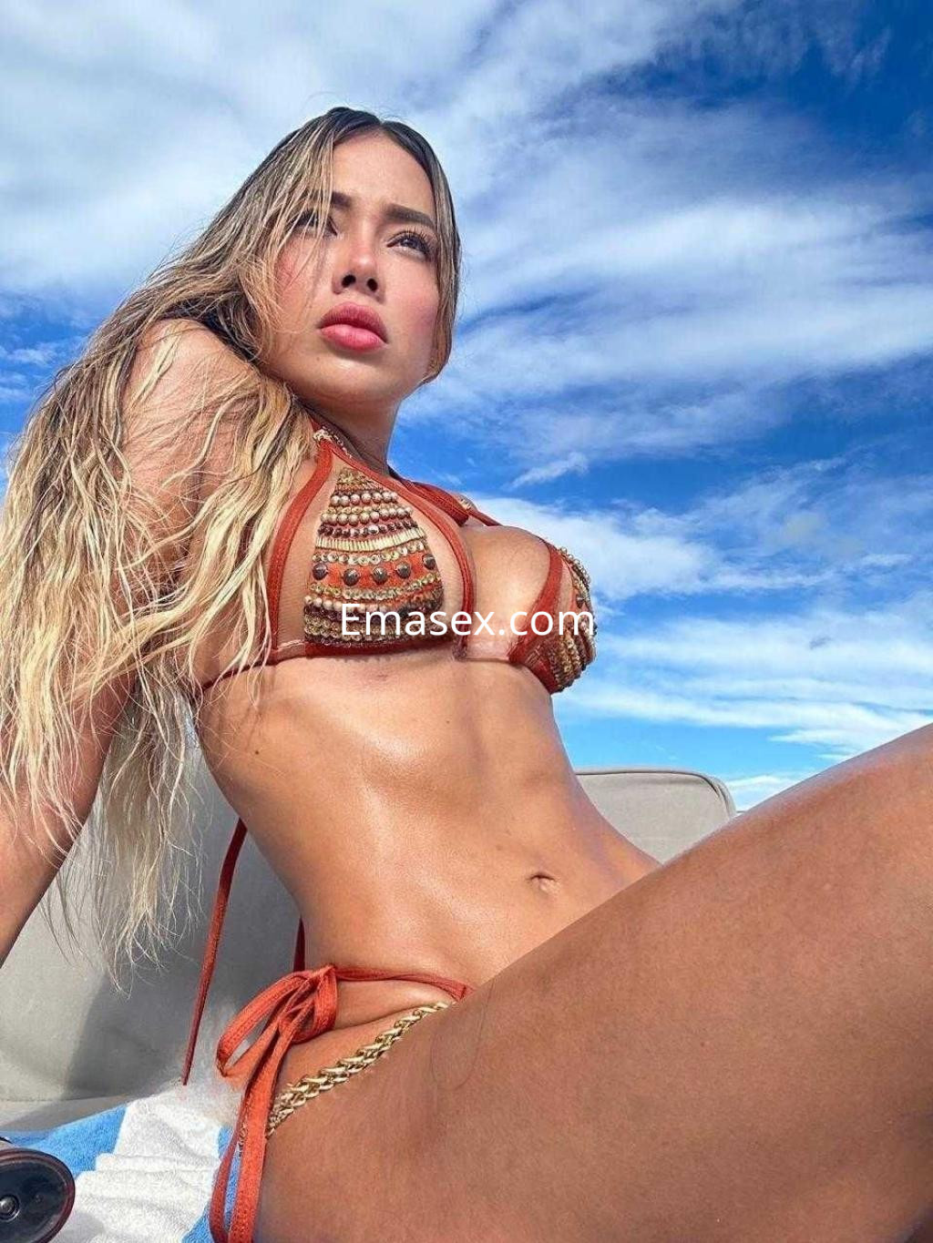 Puta posando en Madrid con un bikini marrón decorado, cabello rubio y piel bronceada bajo un cielo azul.