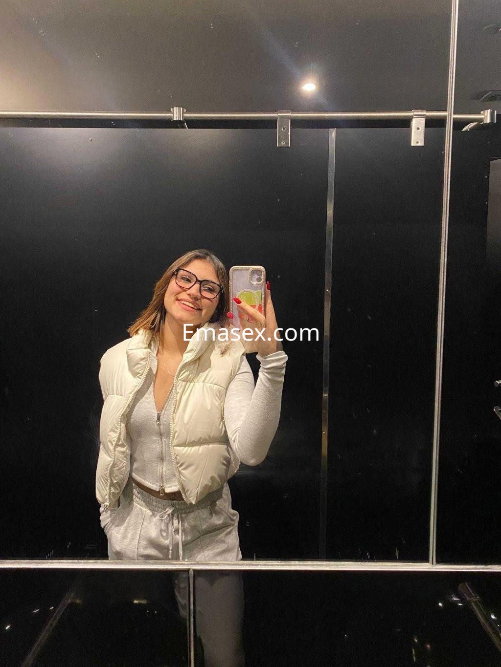 Escort en Murcia sonríe frente al espejo, vistiendo un chaleco blanco y un conjunto de pantalones claros.