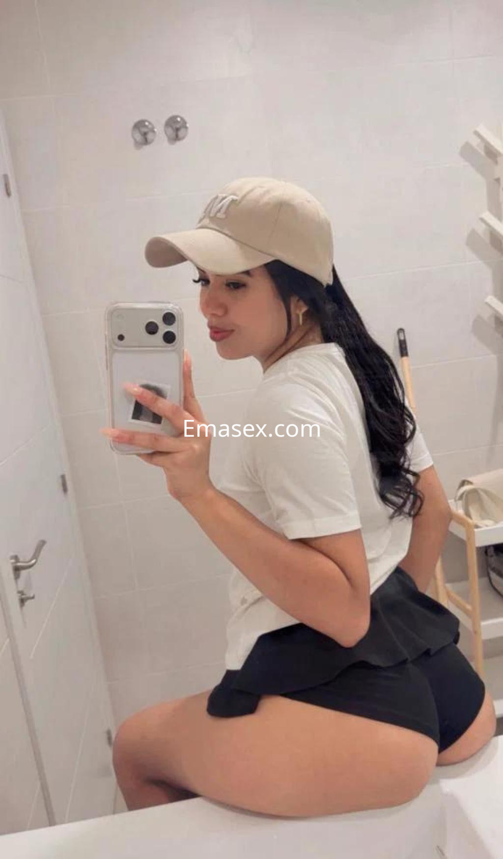 Escort en Murcia con camiseta blanca y gorra, posando en un espejo con un short oscuro.