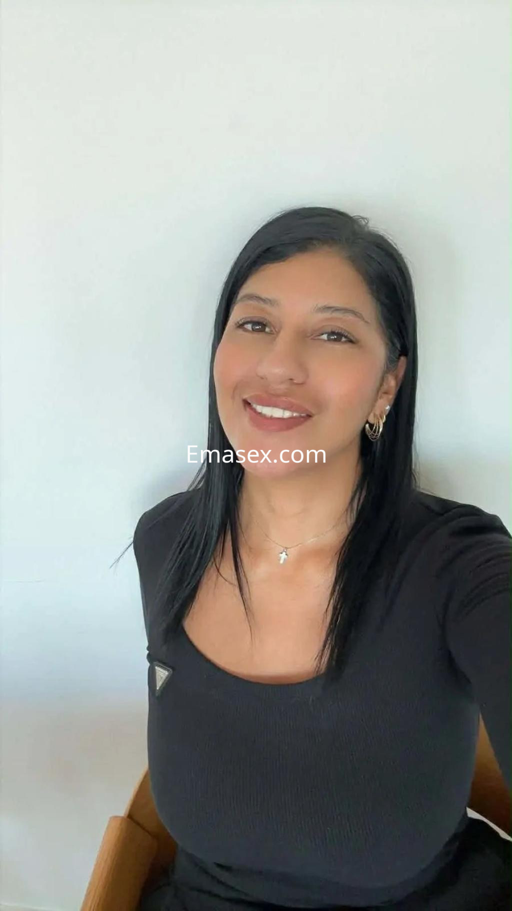 Escort en Murcia con cabello largo y lacio, viste una blusa negra ajustada y sonríe mientras posa sentada.
