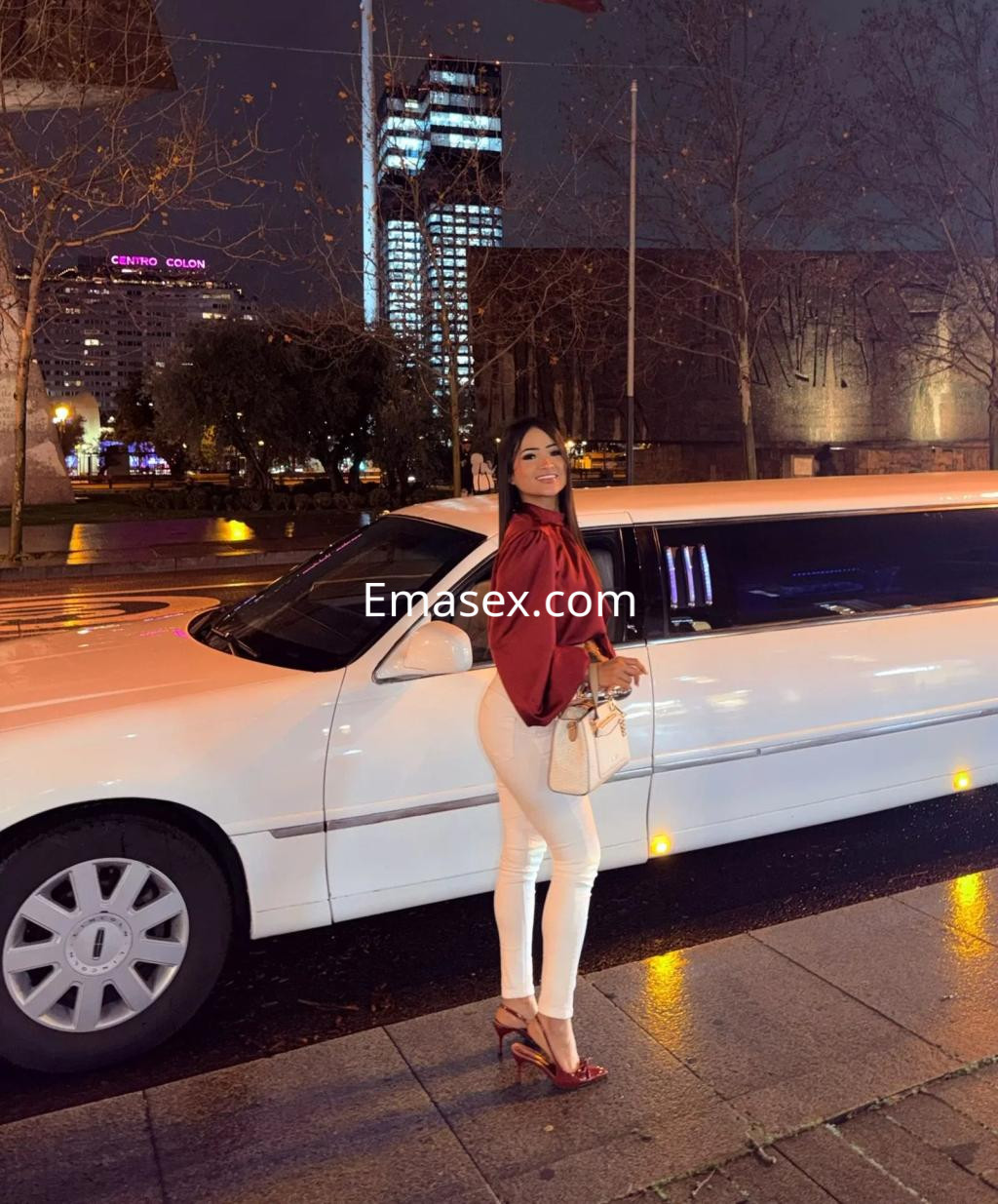 Escort posando junto a una limusina blanca, vestida con blusa roja, pantalones blancos y tacones altos en Murcia.