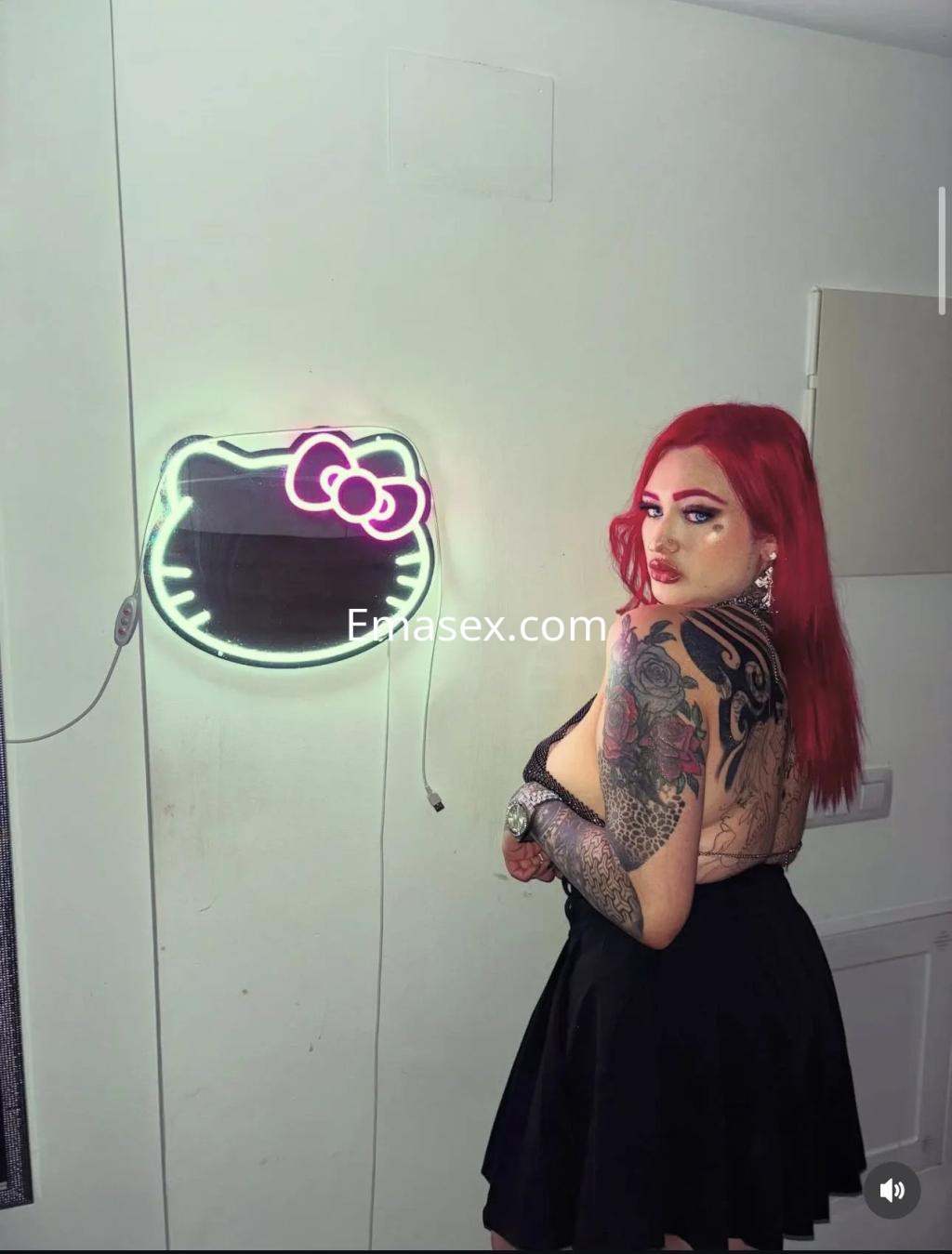 La puta en Murcia muestra un vestido negro con tatuajes visibles y cabello rojo, posando junto a un letrero de neón.