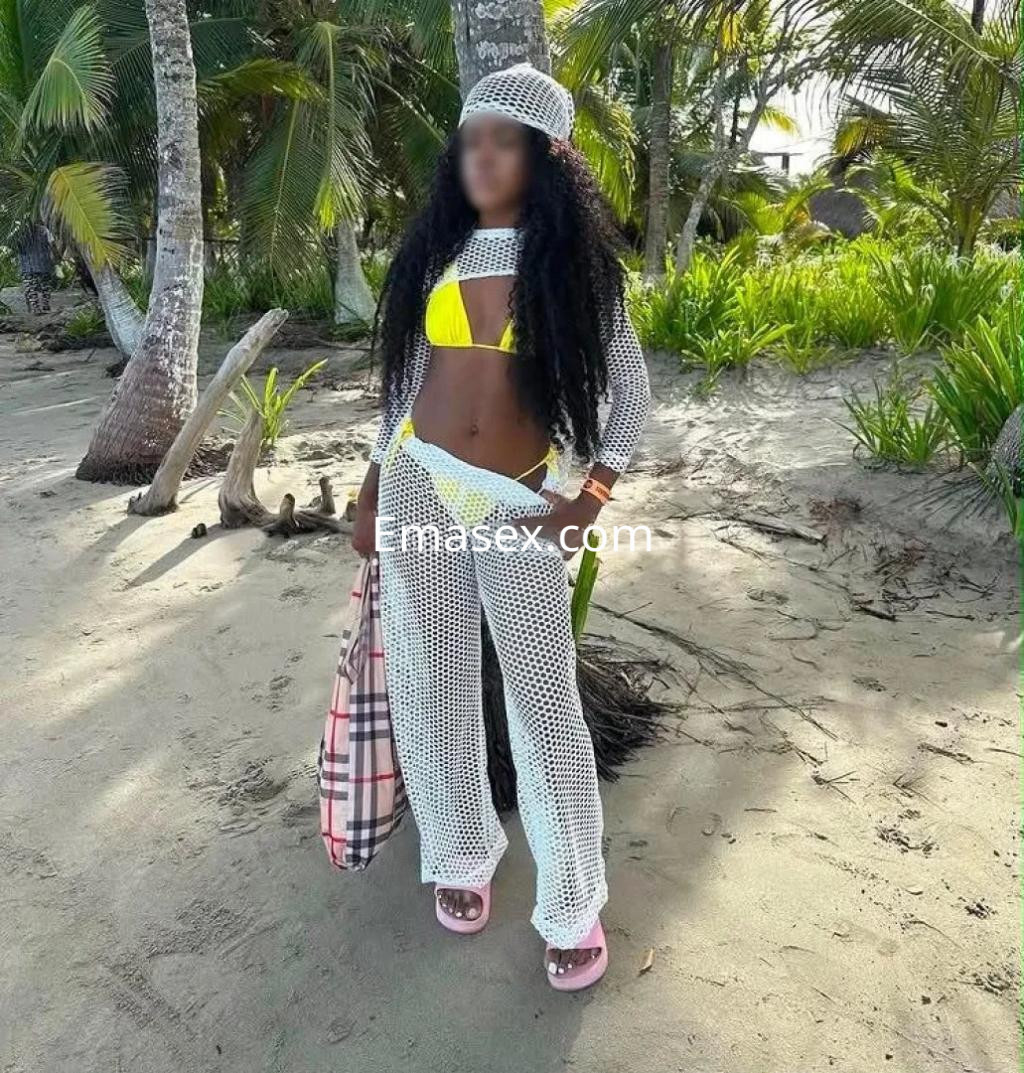 Puta en Gijón con un bikini amarillo y un conjunto de malla blanca, de pie en una playa rodeada de palmeras.