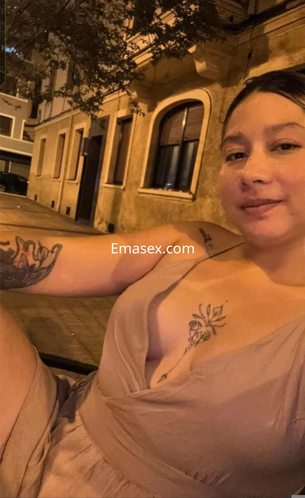 Puta en Oviedo con un vestido claro, mostrando tatuajes en su brazo y una sonrisa relajada en un entorno urbano.