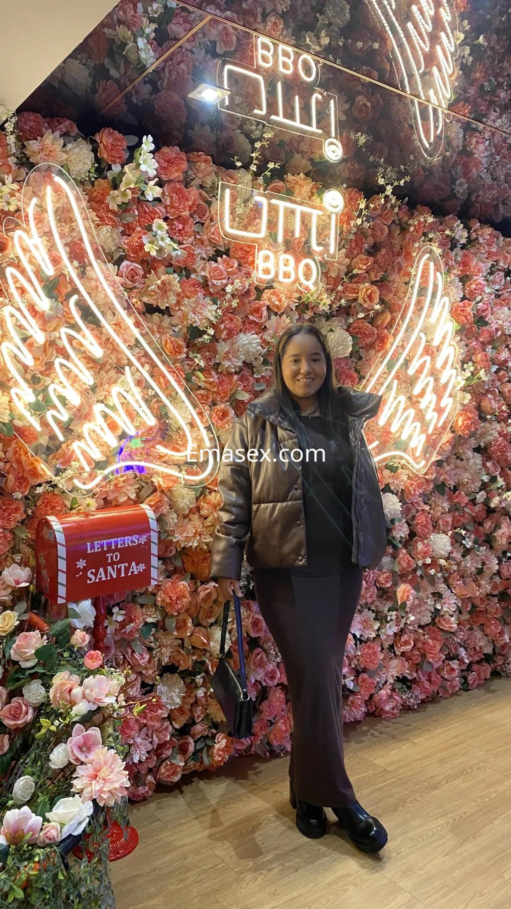 La puta posa con una chaqueta marrón y vestido oscuro frente a un fondo de flores en Vigo.
